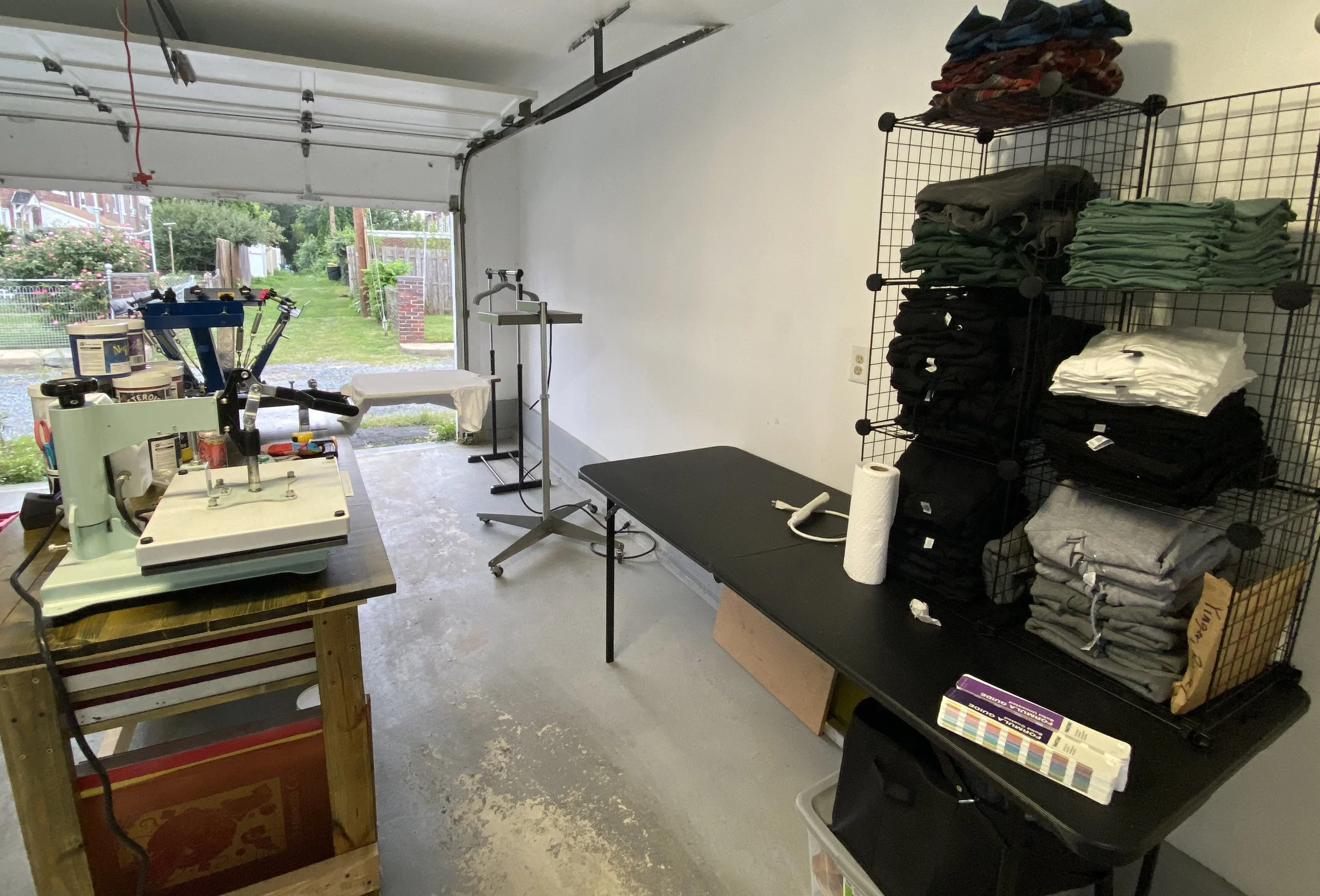 A garage workshop with a table along the wall with stacks of shirts. Another table across from it has a heat press, containers of ink, and a printing press.