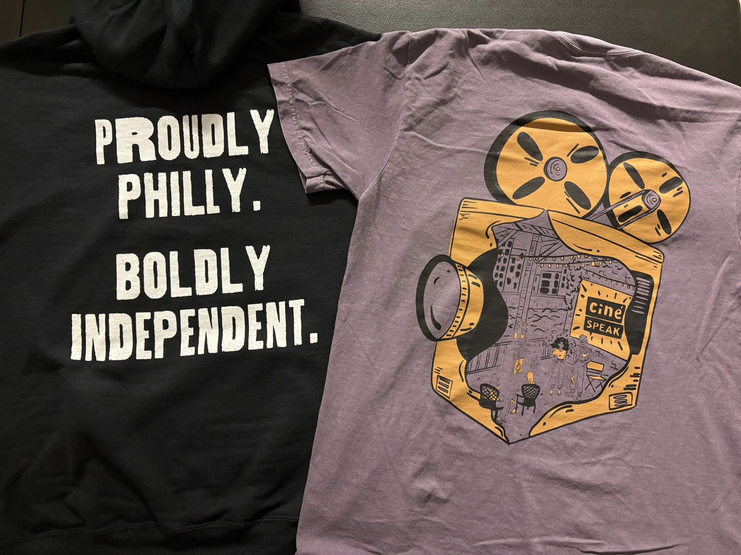 Two T-shirts laid flat. One black with white text that reads 'PROUDLY PHILLY. BOLDLY INDEPENDENT.' The other purple with a colorful graphic of a movie camera with an illustration of a movie screen and people watching it inside of the camera