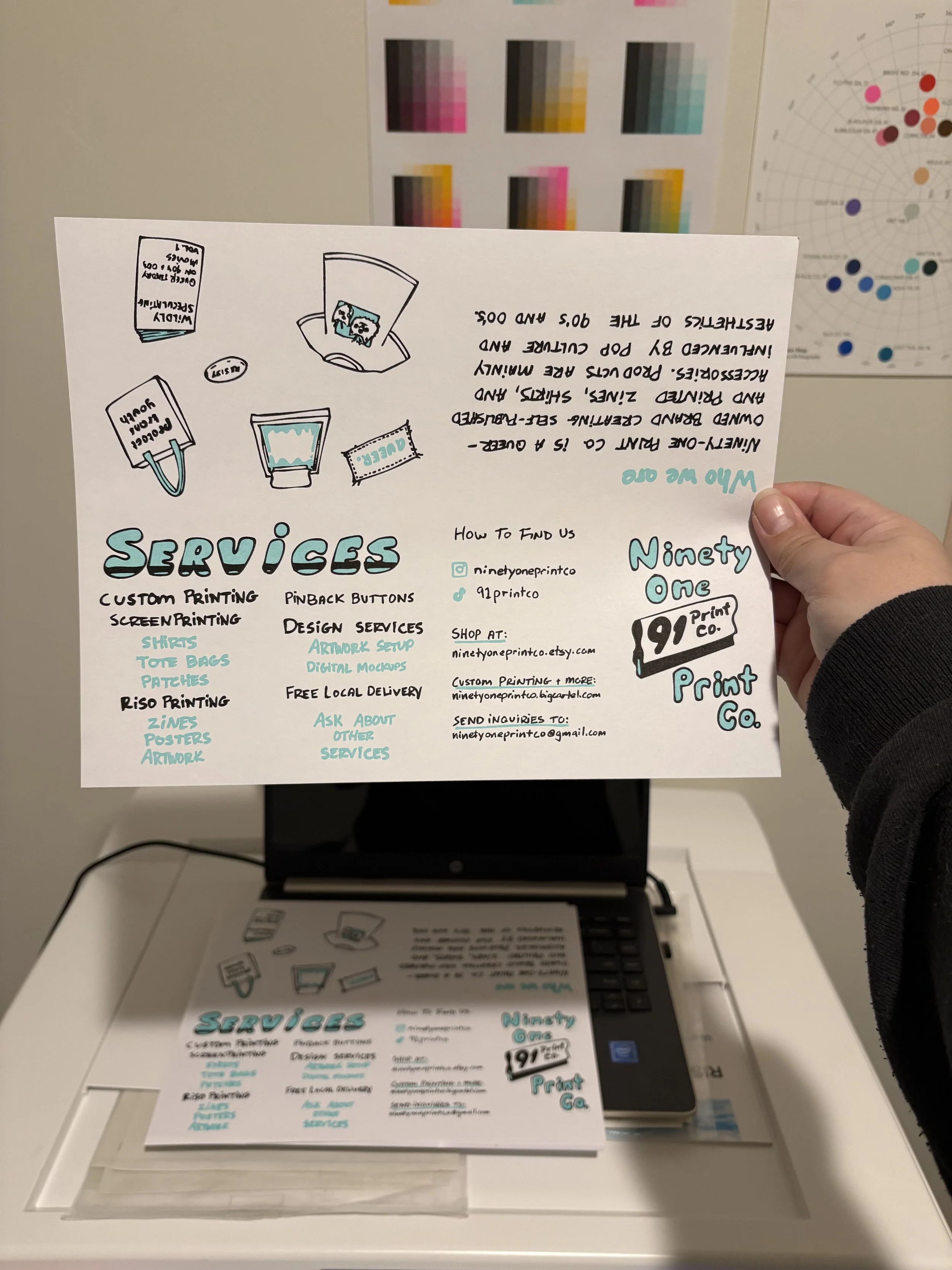 A risograph printed one-page zine, unfolded, held in a hand, about services offered by Ninety-One Print Co. The colors used to print the zine are mint and black. Behind the zine is a stack of more prints on top of a laptop on top of a RISO machine