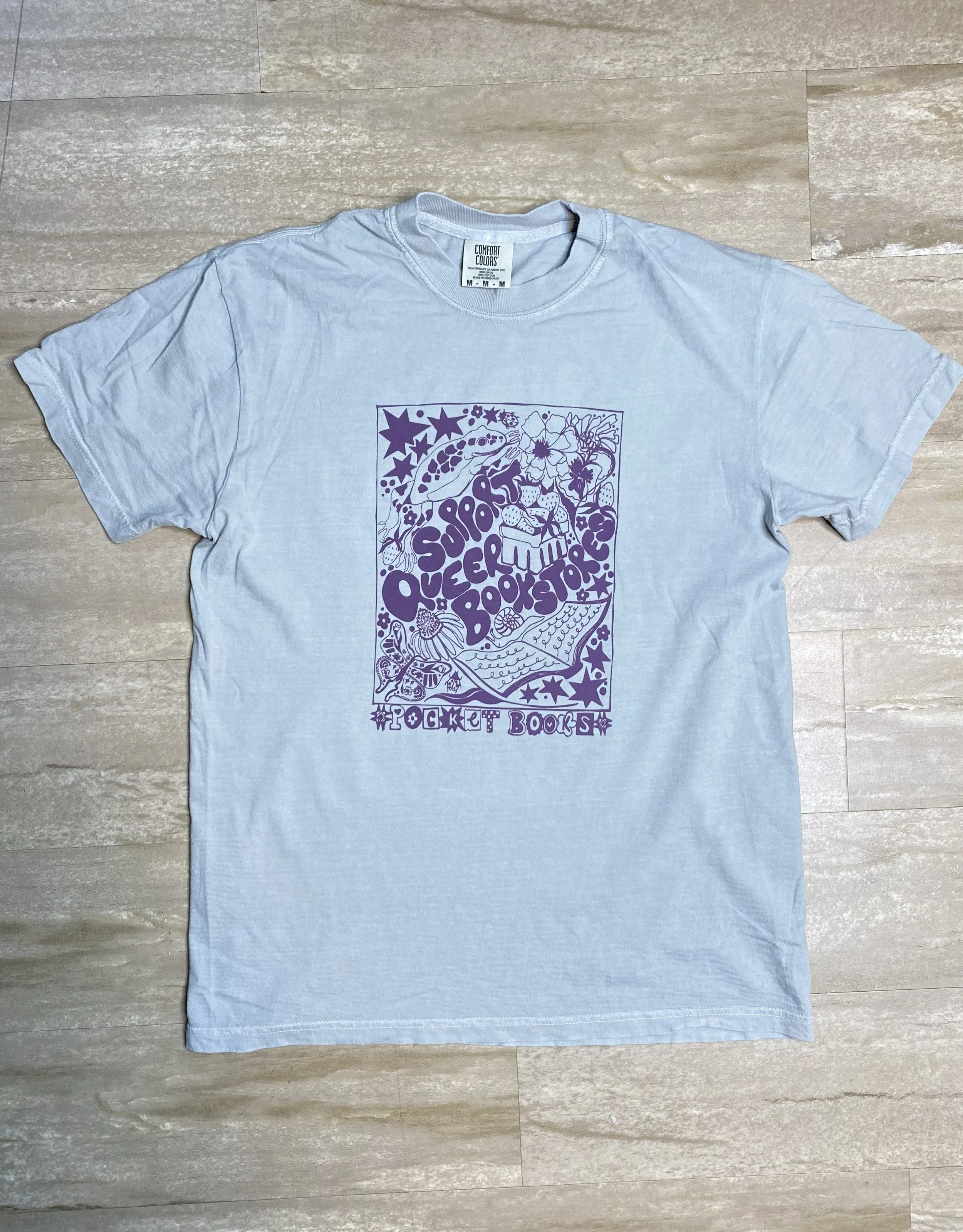 A light grayish teal T-shirt with a purple graphic design on the front that includes the text 'Pocket Books' and 'Support Queer Bookstores" along with illustrations of a book, a frog, strawberries, a butterfly, and other small deatils