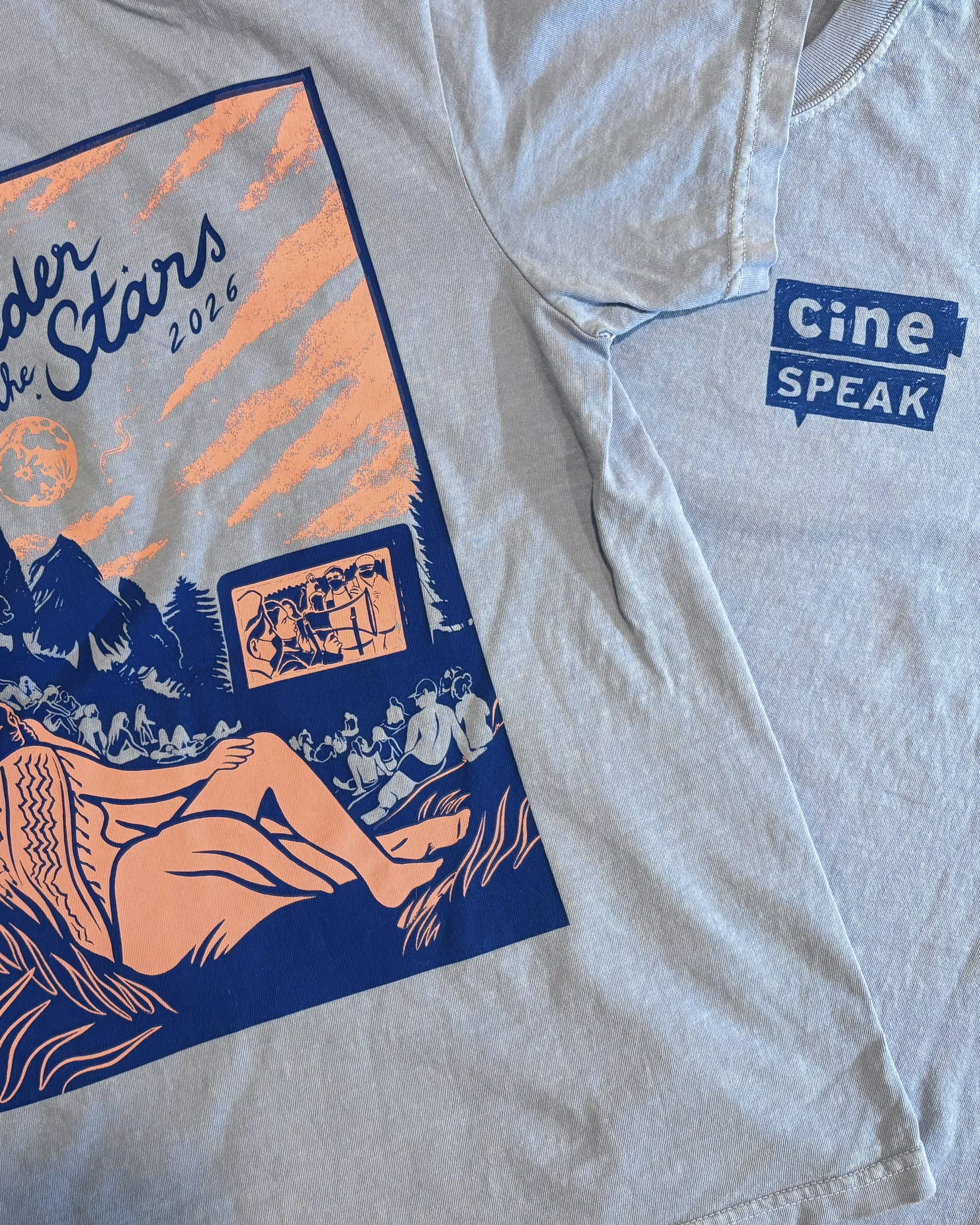 A light blue T-shirt with a blue and orange graphic design featuring trees and people looking at a movie screen.