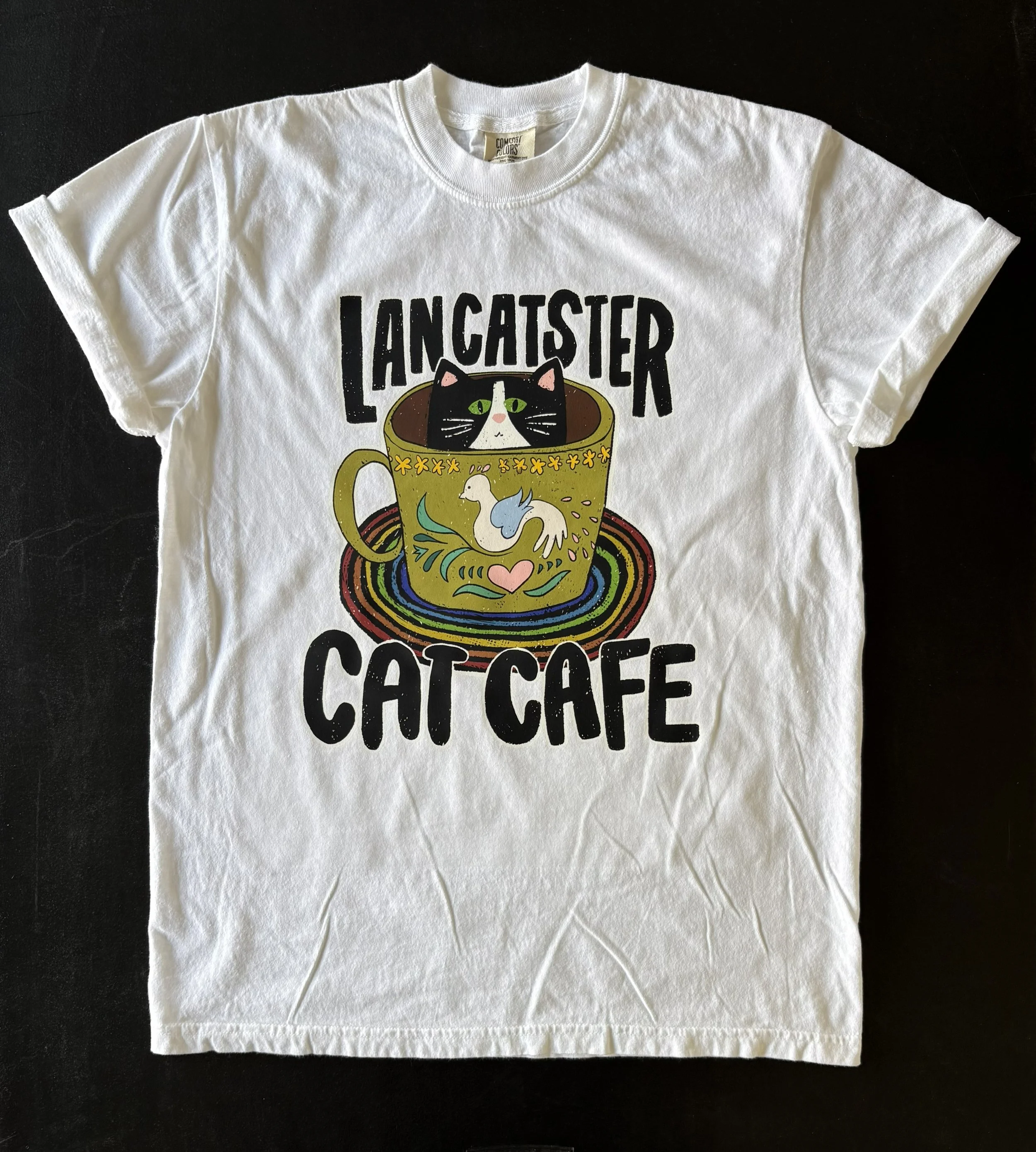 white T-shirt with the words "Lancatster Cat Cafe" printed on it, featuring a colorful illustration of a black and white cat inside a coffee cup with a rainbow swirl pattern.