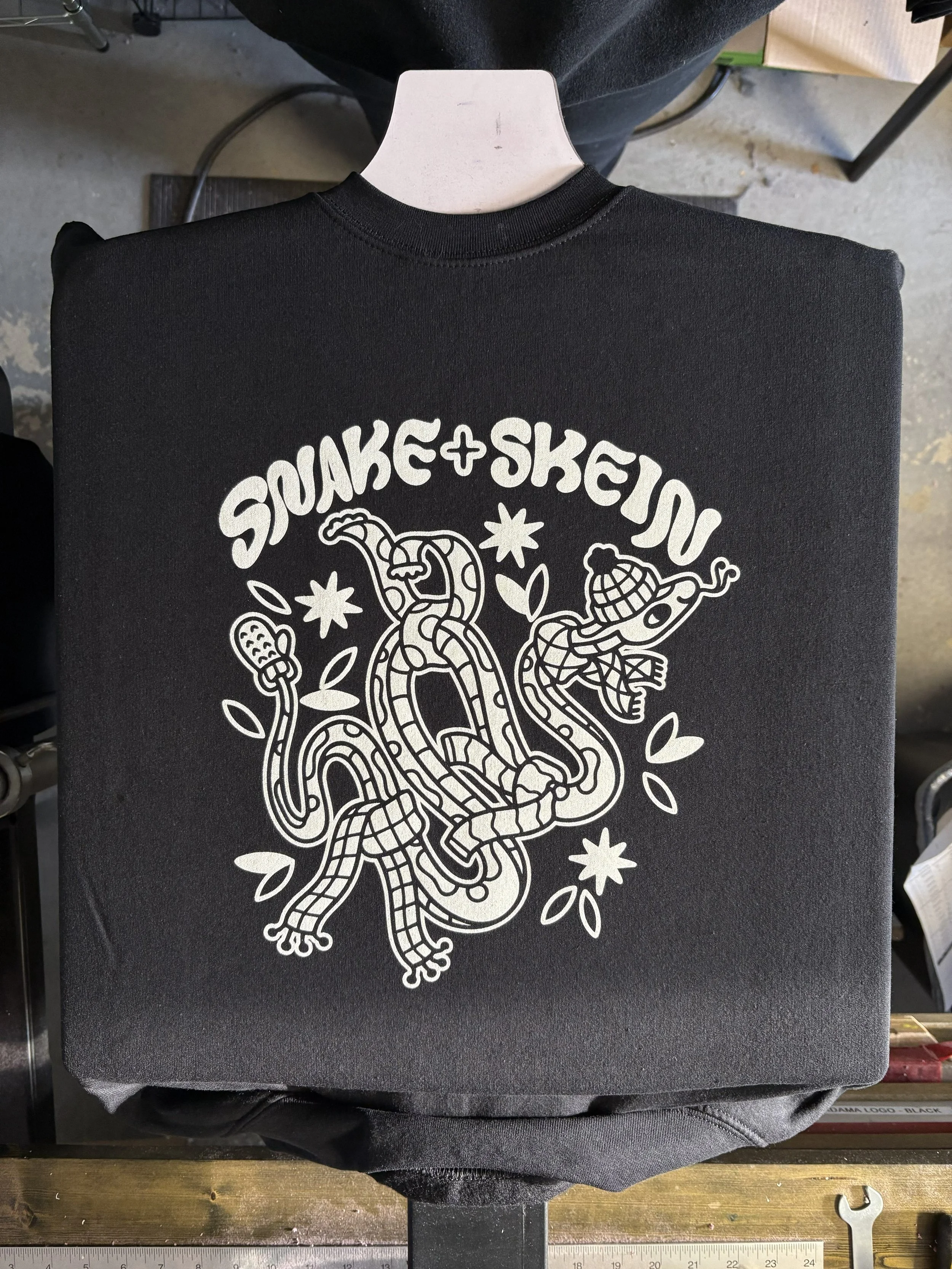 Black shirt on a printing press with a white design featuring a snake wearing a scarf and hat. Above the snake is the text "Snake + Skein"