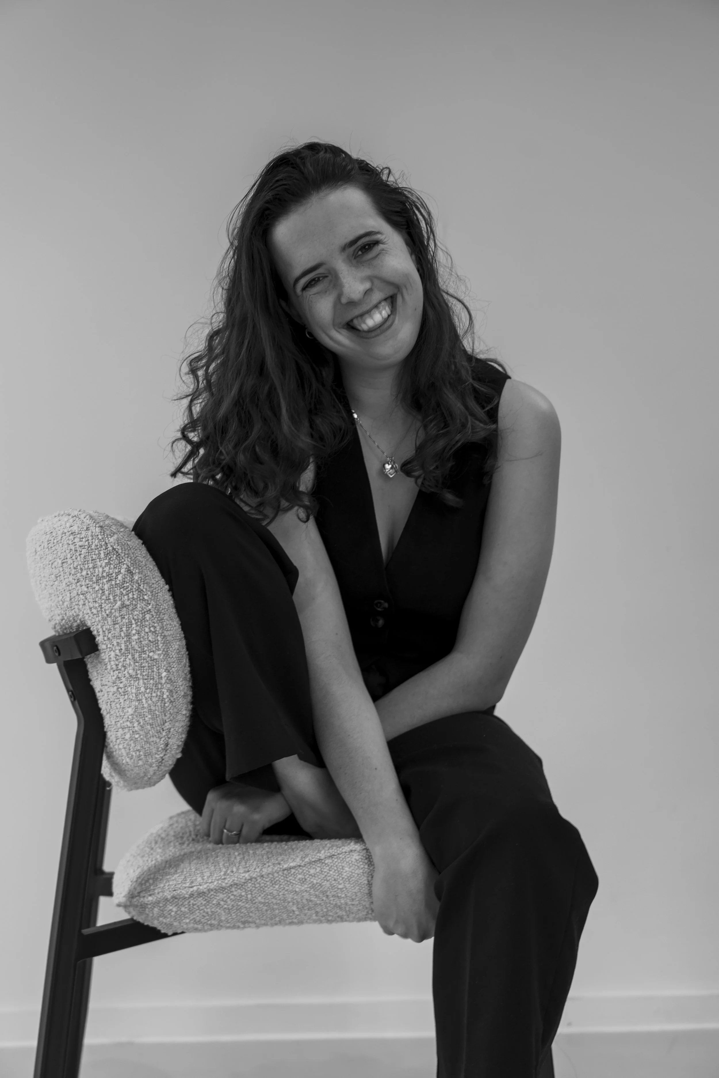 Zwarte en witte foto van een lachende vrouw met donker haar, gekleed in een zwart top en zwart broek, zittend op een stoel met een lichte armleuning, tegen een effen achtergrond.