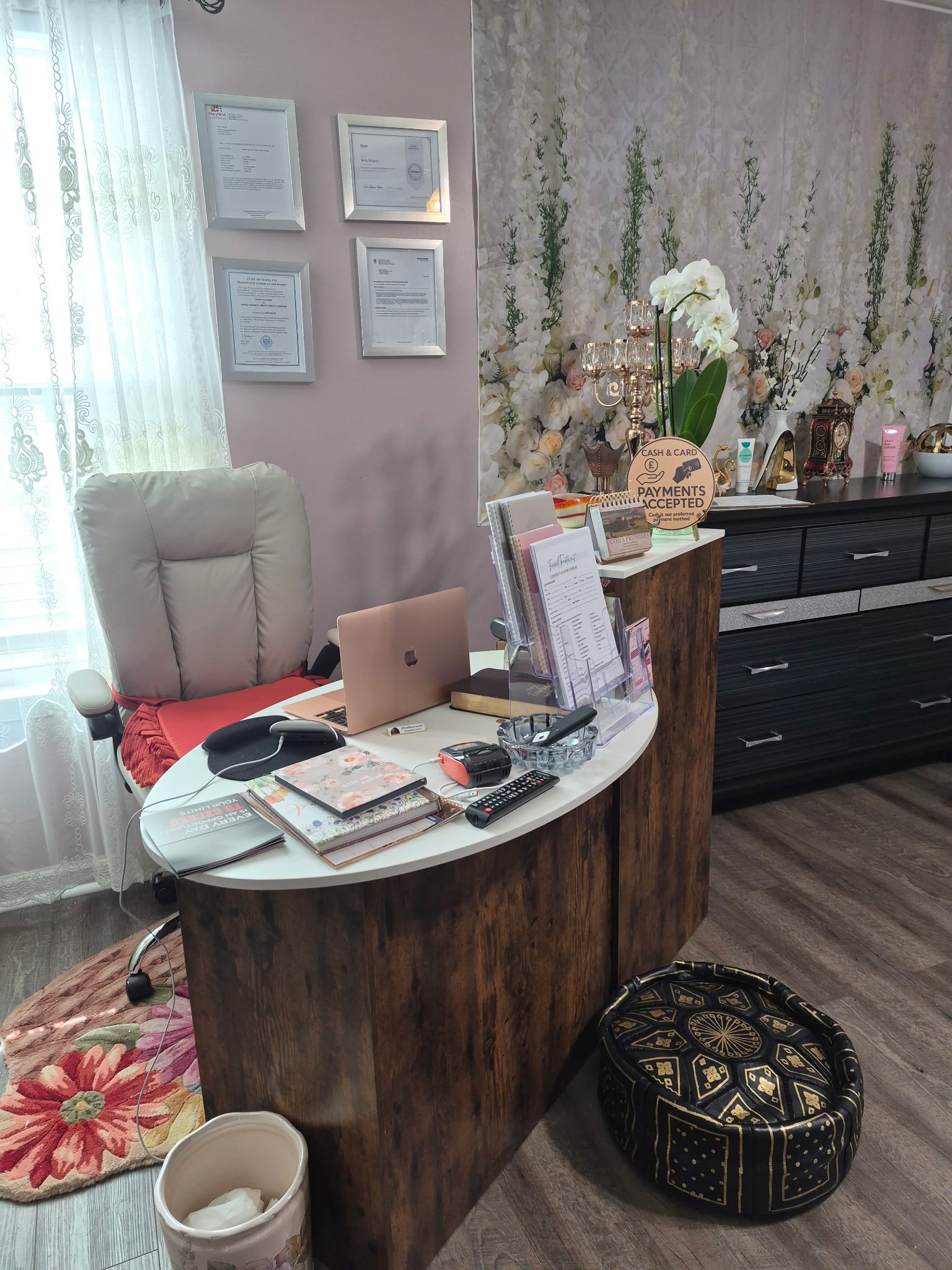 Office with a white and wood desk, a beige chair, a floral rug, and various items including notebooks, a laptop, and stationery. A dark wood cabinet with decorative objects and flowers. Framed certificates on the wall, a floral wallpaper, and light-filtering curtains.