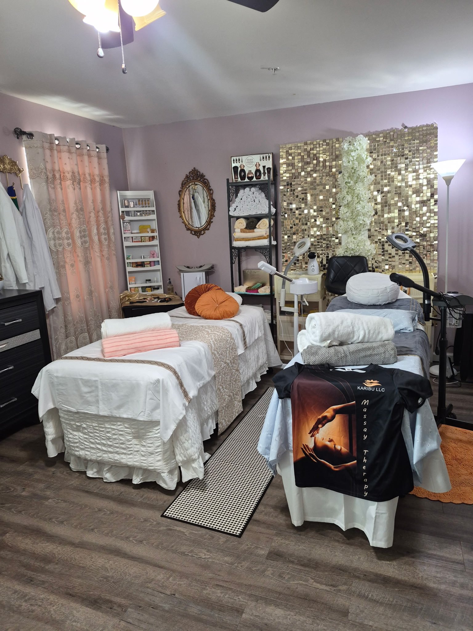 Room with multiple beds, towels, and equipment associated with wellness or spa treatments. The room has lavender-colored walls, a gold mirror, and decorative wall panels with flowers and reflective tiles. Towels and supplies are organized on shelves and tables, and there are lamps and lighting fixtures.