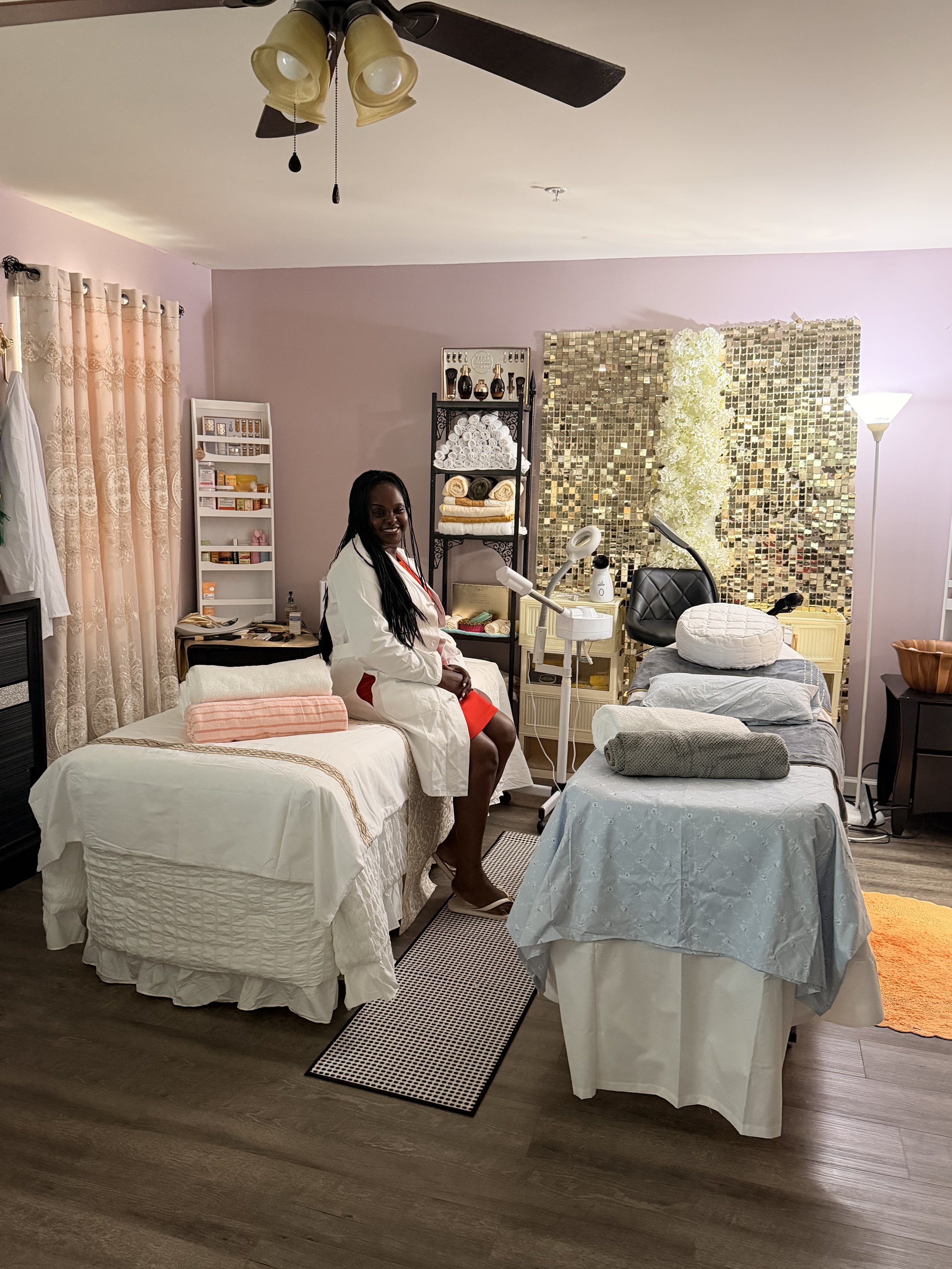 CEO sitting on a bed in a room with spa or massage equipment, with towels, skincare products, and lamps around. The room has pink and purple walls, a gold-tiled accent wall, and wooden flooring.
