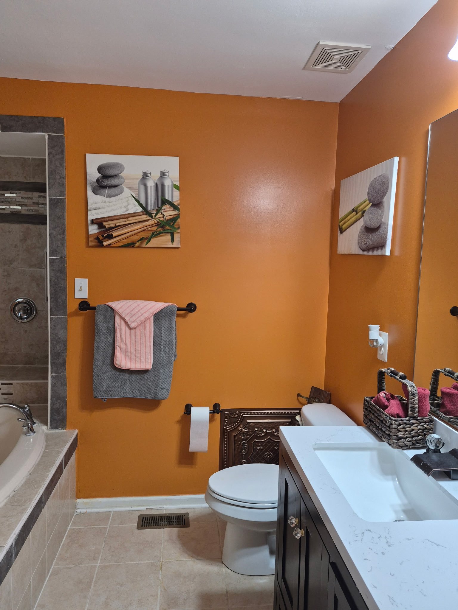 A bathroom with a white countertop sink, dark wooden cabinet, toilet, and orange walls. There are two pieces of art on the wall, a towel hanging on a black towel bar, a basket with pink towels, and a mirror.
