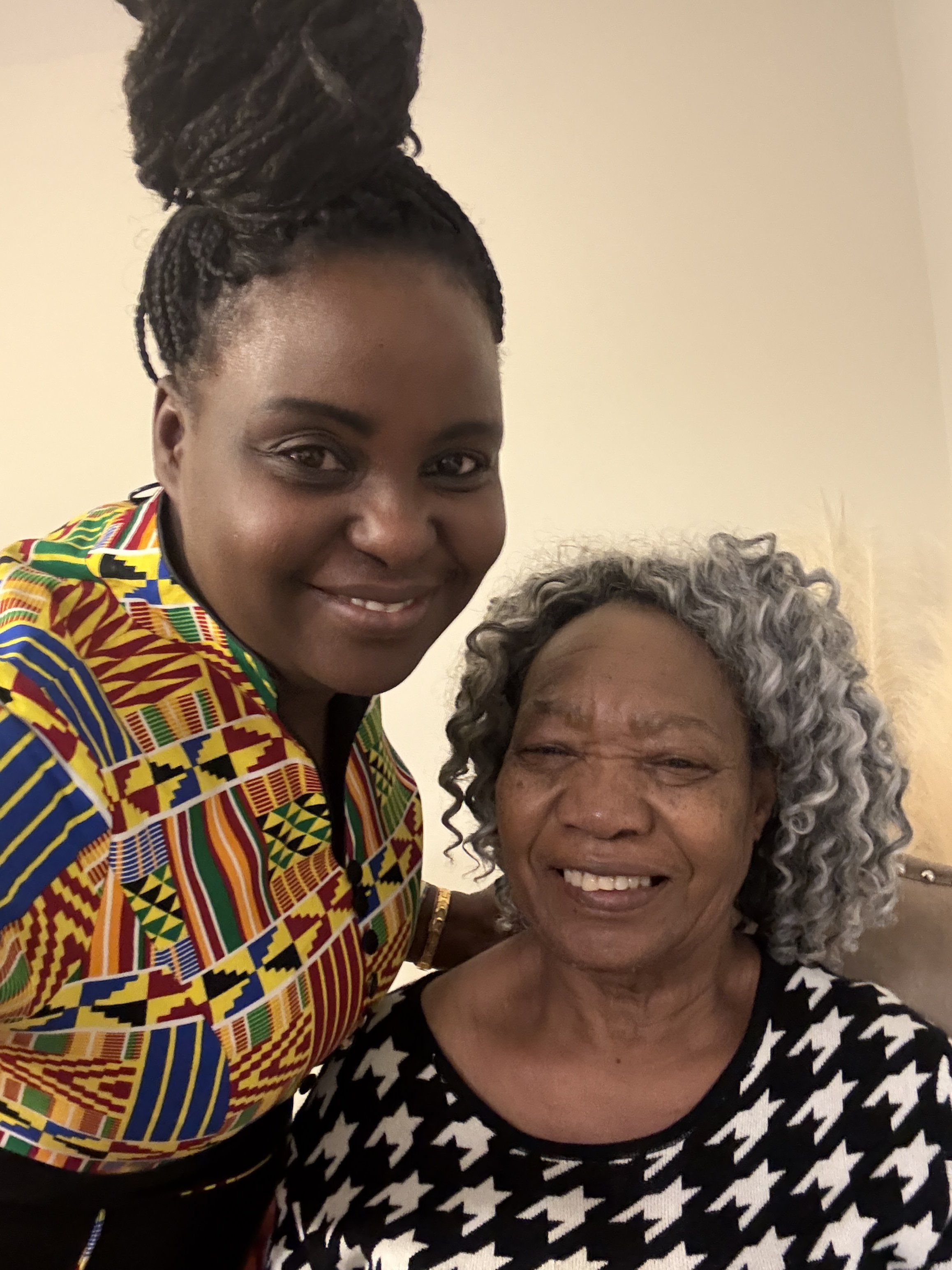 A woman wearing a colorful African print shirt smiling and taking a selfie with an older woman with curly gray hair, who is also smiling.