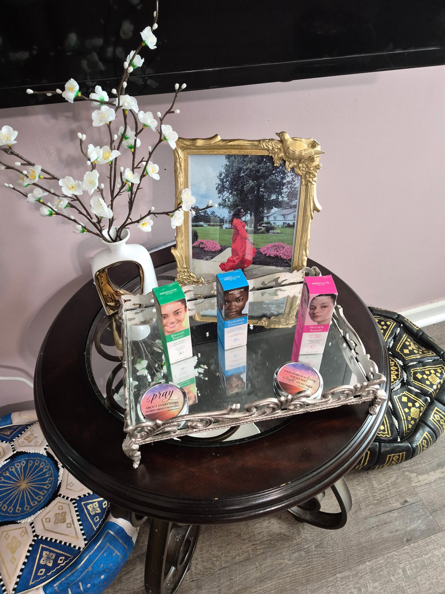 Decorative table with makeup products, a vase with artificial white flowers, a picture frame, and a decorative tray with motivational quotes.