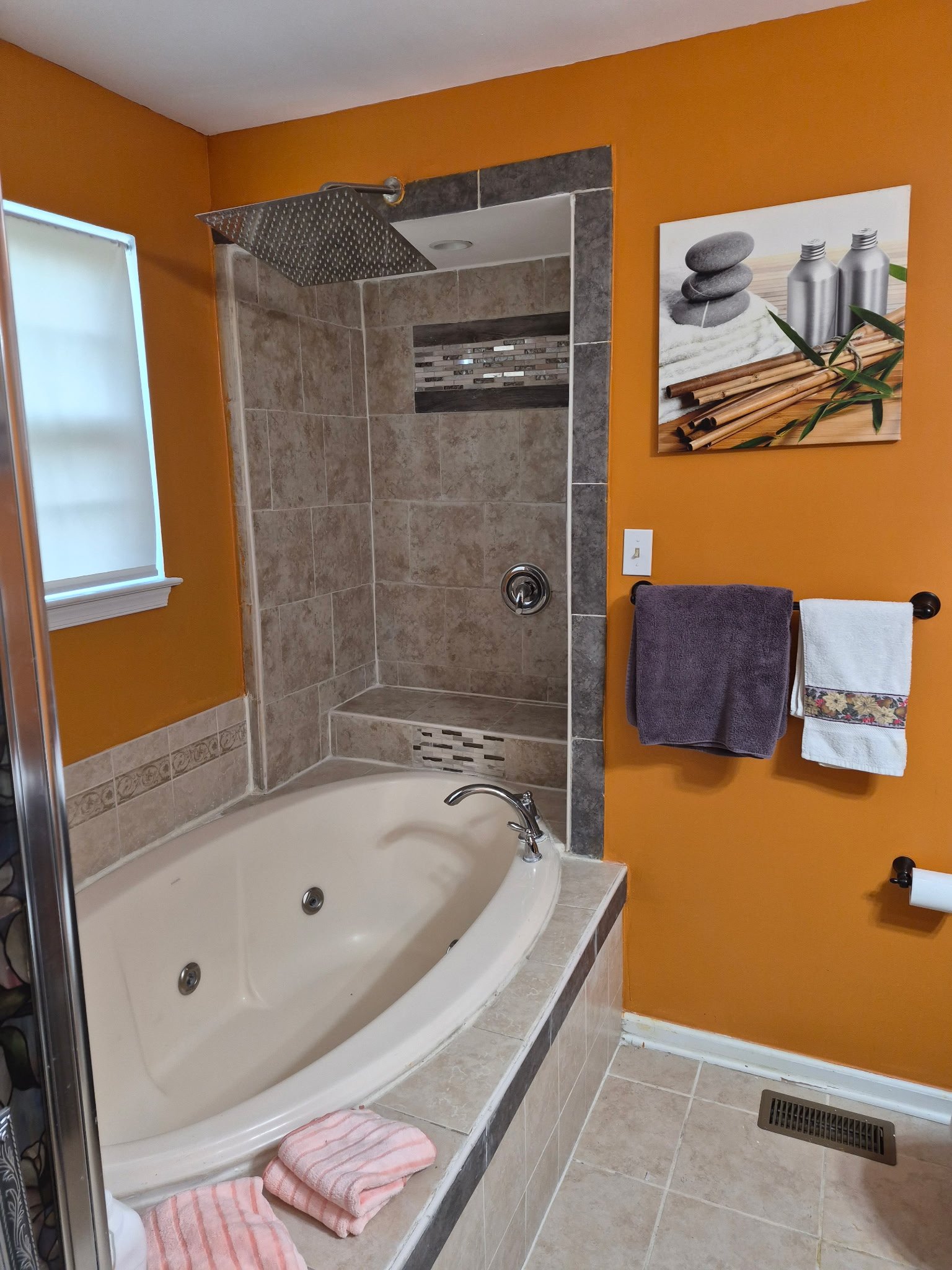 A bathroom with an orange wall, featuring a bathtub, a tiled shower area, and a decorative wall art of stacked stones and bottles. Towels are hanging on a rack, and rolled towels are placed on the bathtub's edge near a pink and white striped rug.