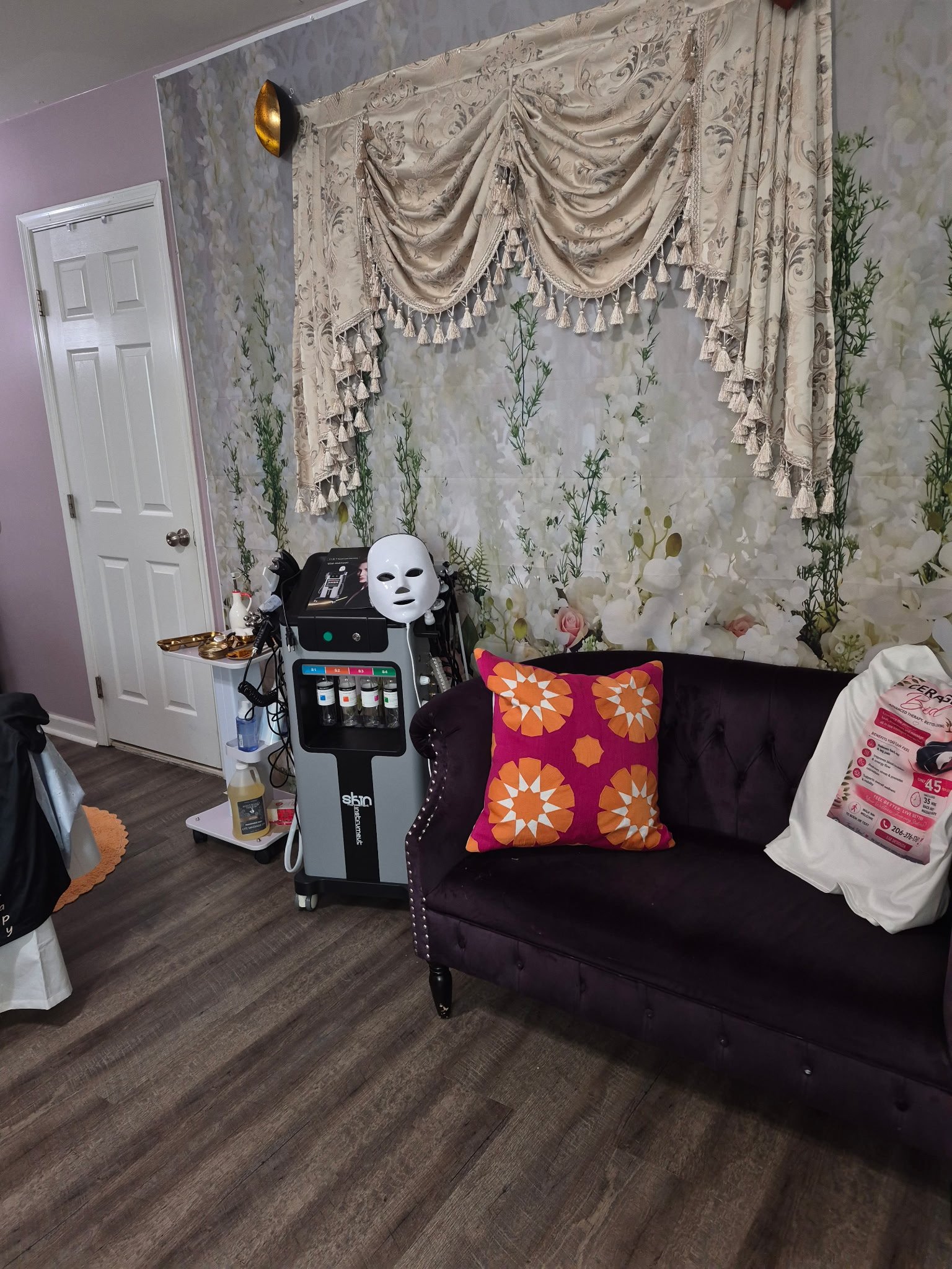 A salon or spa treatment room with a floral patterned wall, a velvet purple sofa with orange and pink patterned pillow, a white door, and a treatment machine with a white mask and various bottles on a cart.