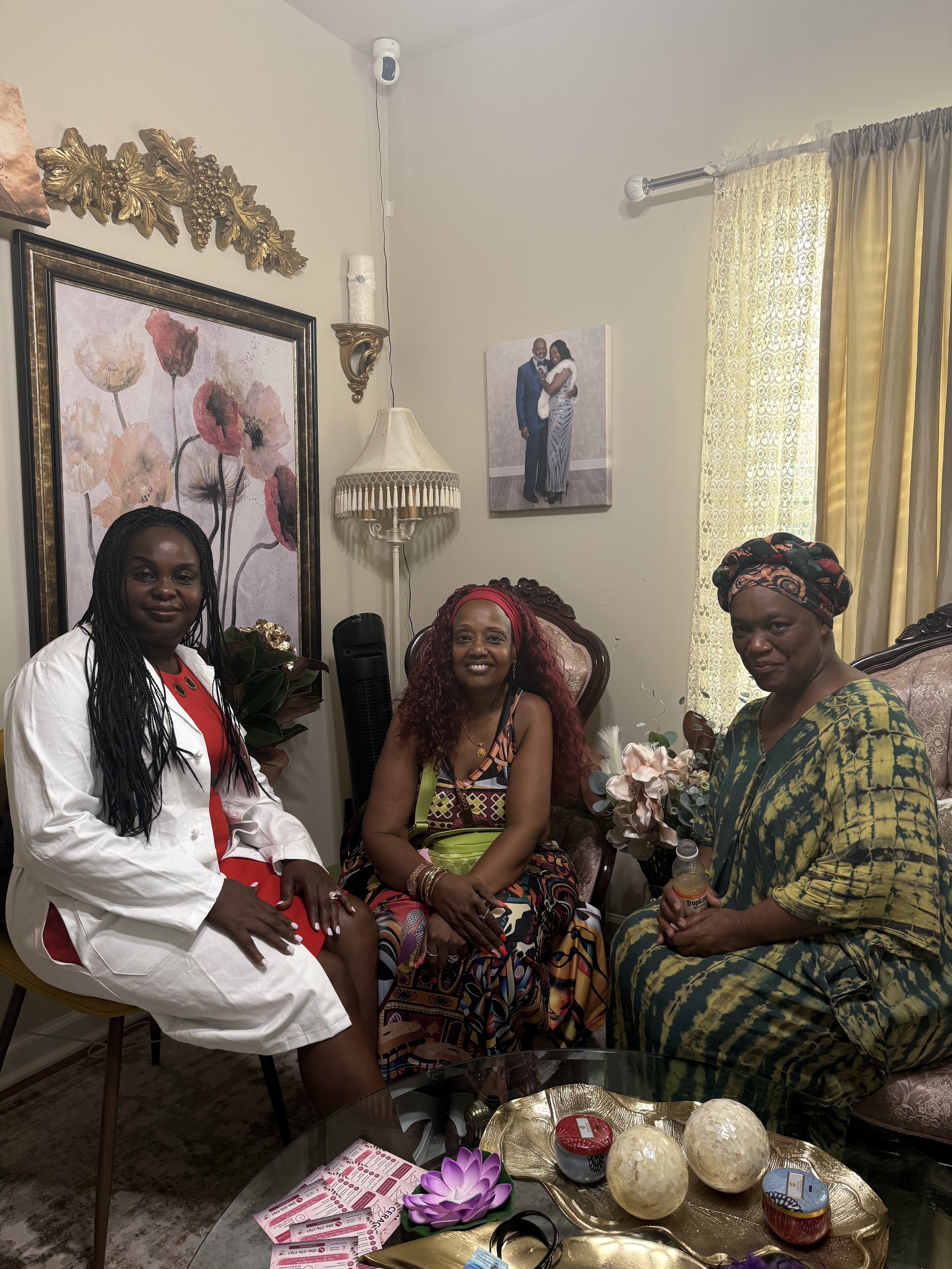 Three women sitting and standing around a table in a cozy living room, with a floral painting, a framed wedding photo, and decorative lamps on the wall.