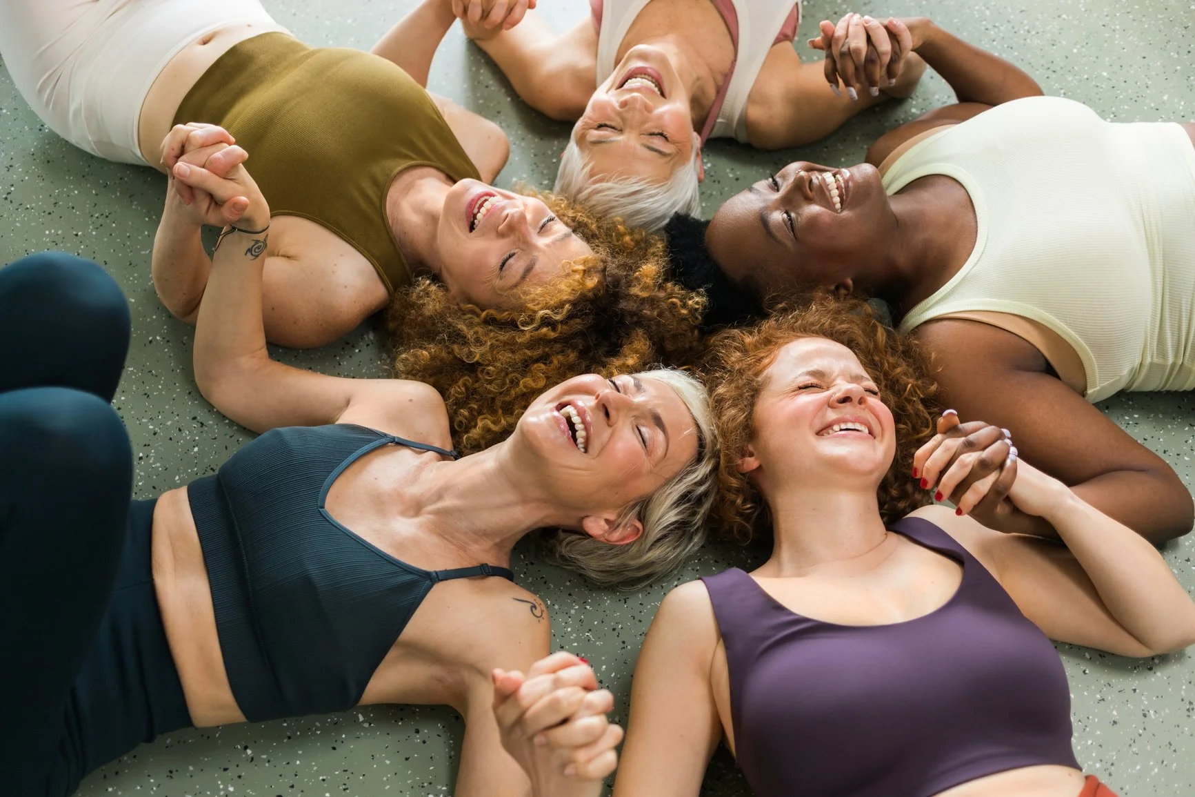 Six women lying on the gym floor in a circle holding hands and smiling.