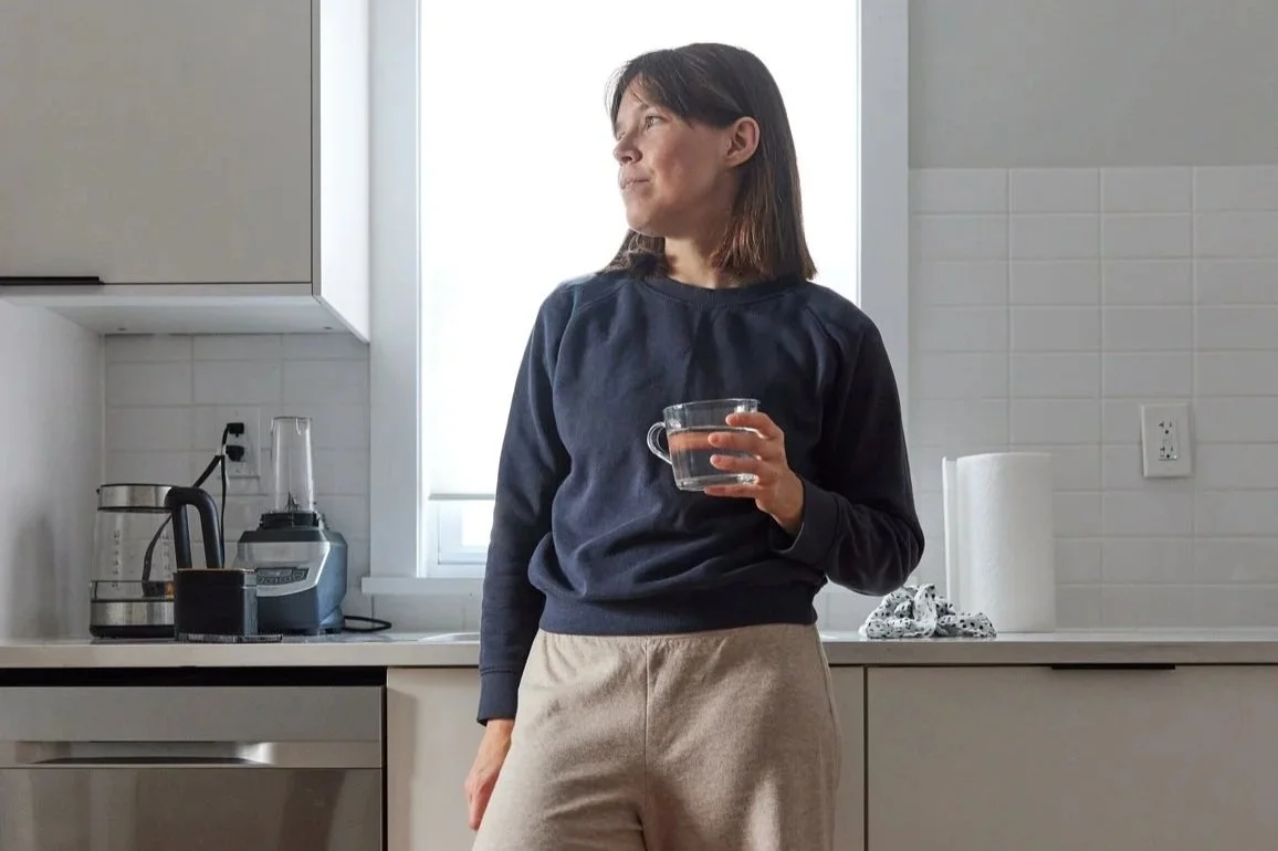 Woman standing in kitchen holding a glass mug, looking thoughtfully out the window.