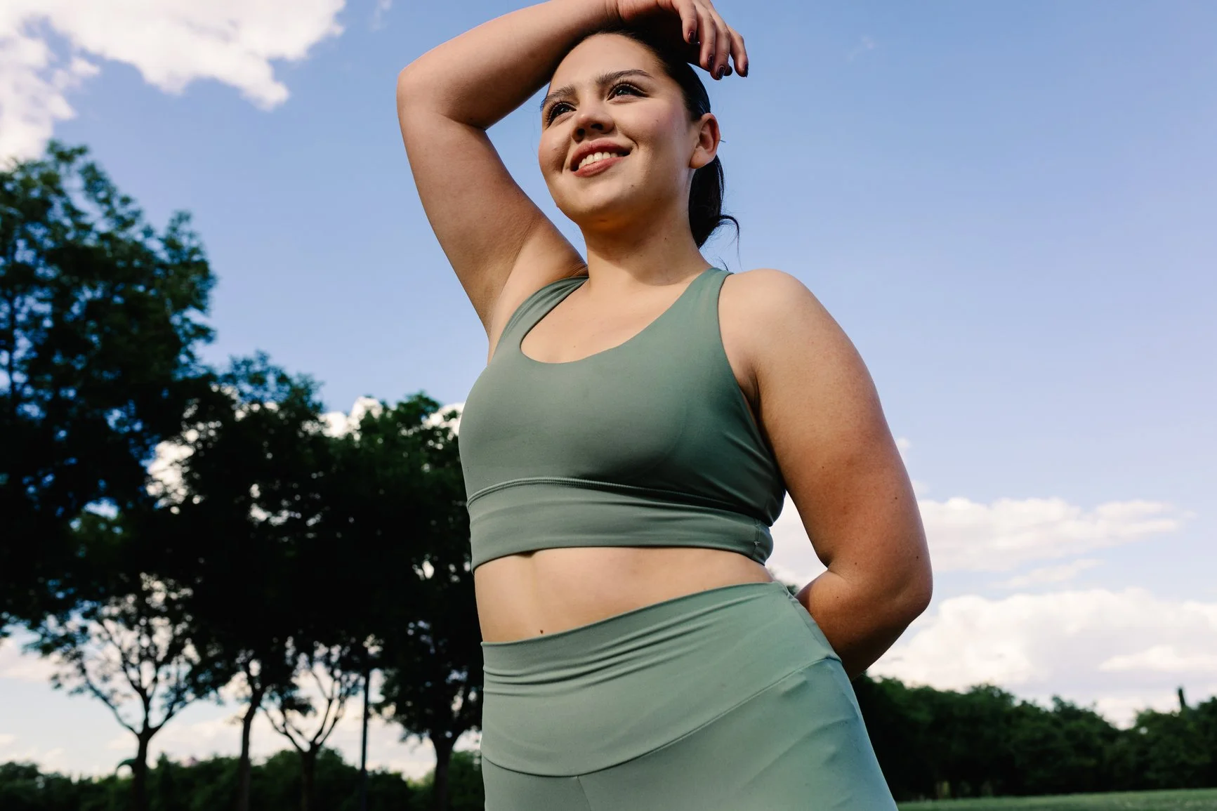 A woman in green athletic wear smiling outdoors with one hand on her head, trees and a blue sky with clouds in the background.