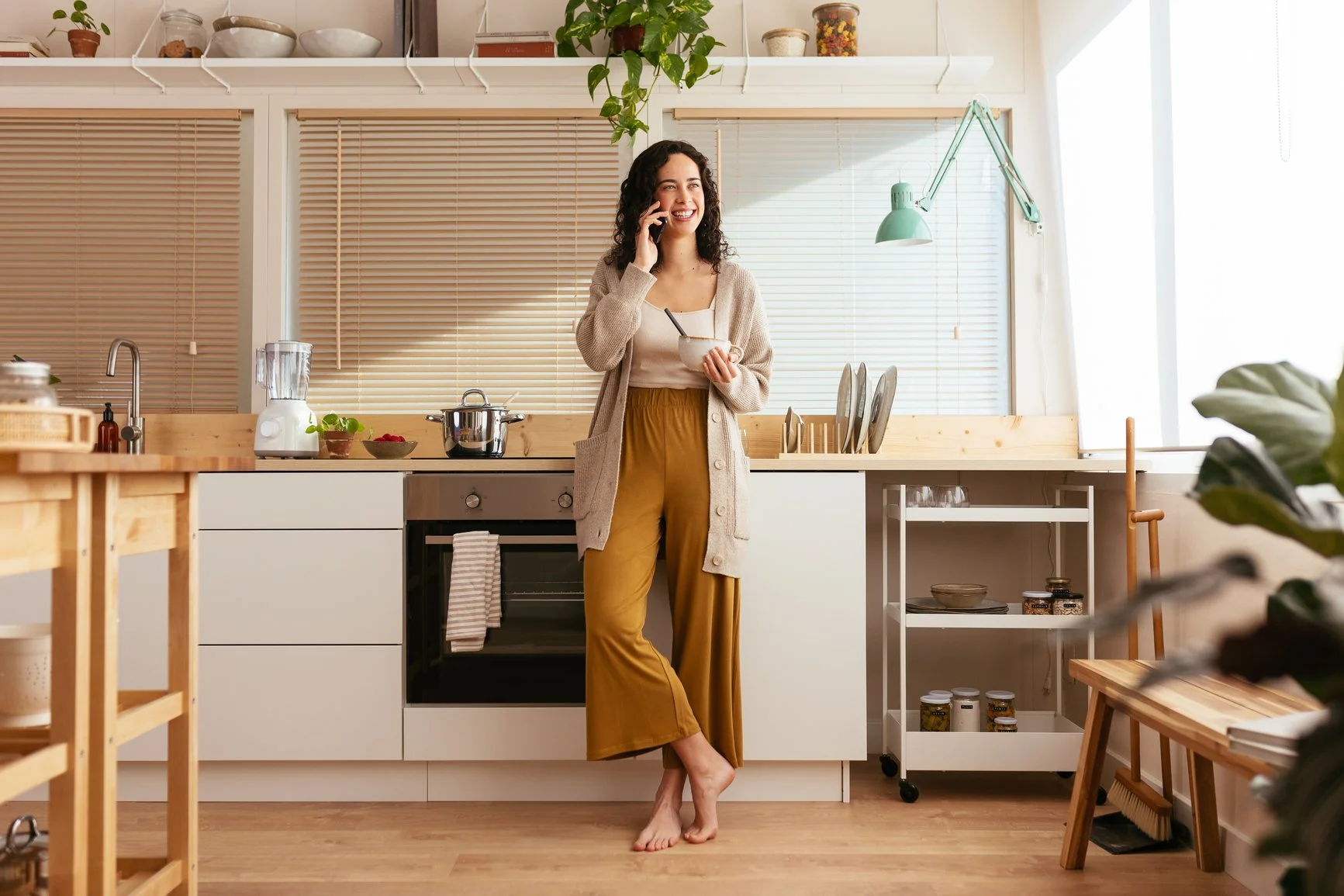 A woman standing in a modern kitchen, talking on a phone, holding a mug, while smiling. The kitchen has white cabinets, wooden accents, and is well-lit with natural light. She is barefoot, wearing mustard-colored pants and a beige cardigan.