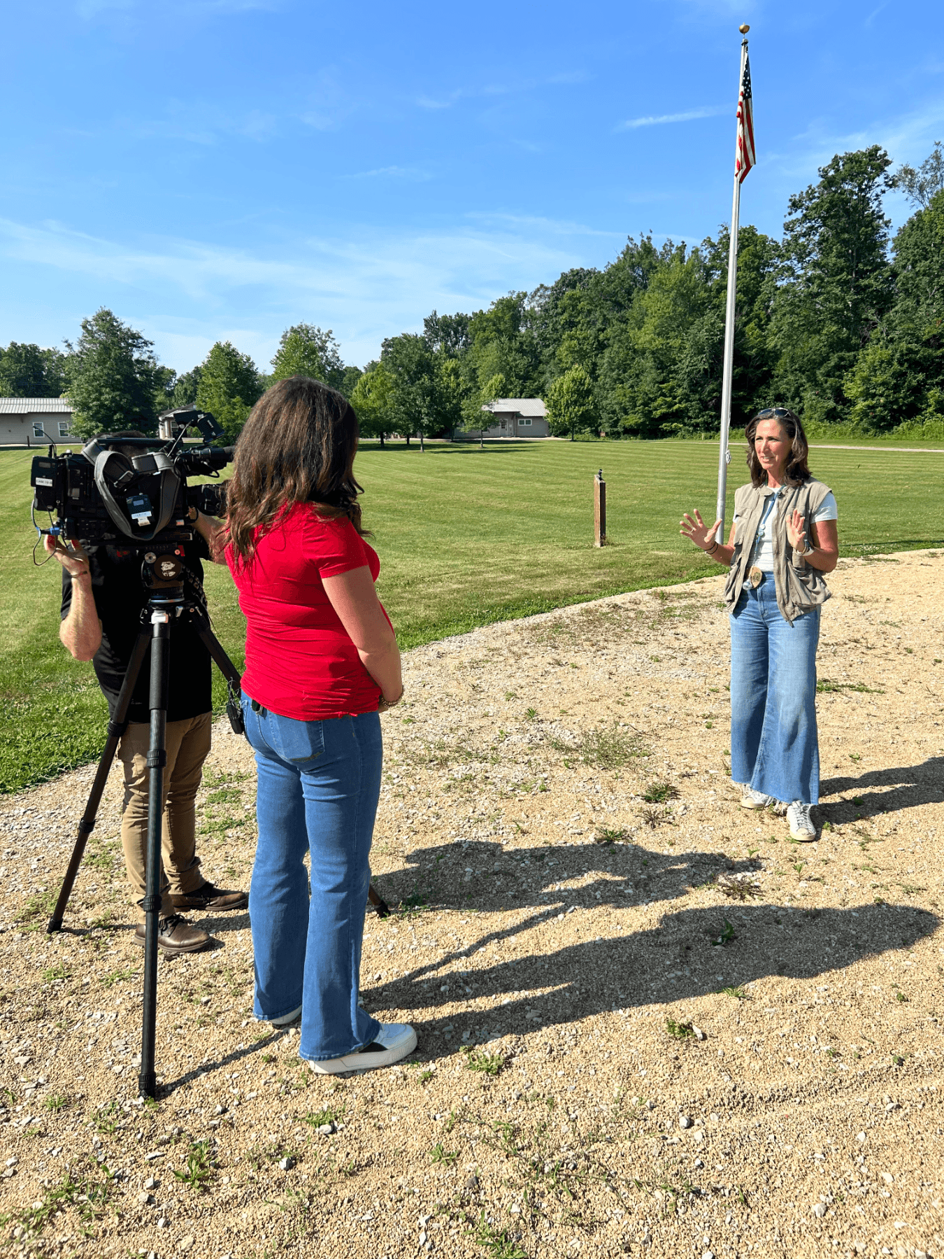 A woman is speaking outdoors near a flagpole with an American flag. Two other people are filming her, one with a professional video camera. The scene takes place in a grassy, open area with trees and some buildings in the background on a sunny day.