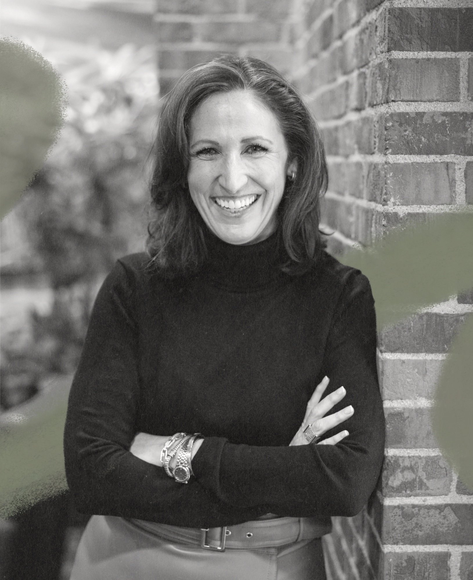 Woman smiling, with brown hair, wearing a black turtleneck, accessorized with watches and bracelets on her left wrist, standing against a brick wall.