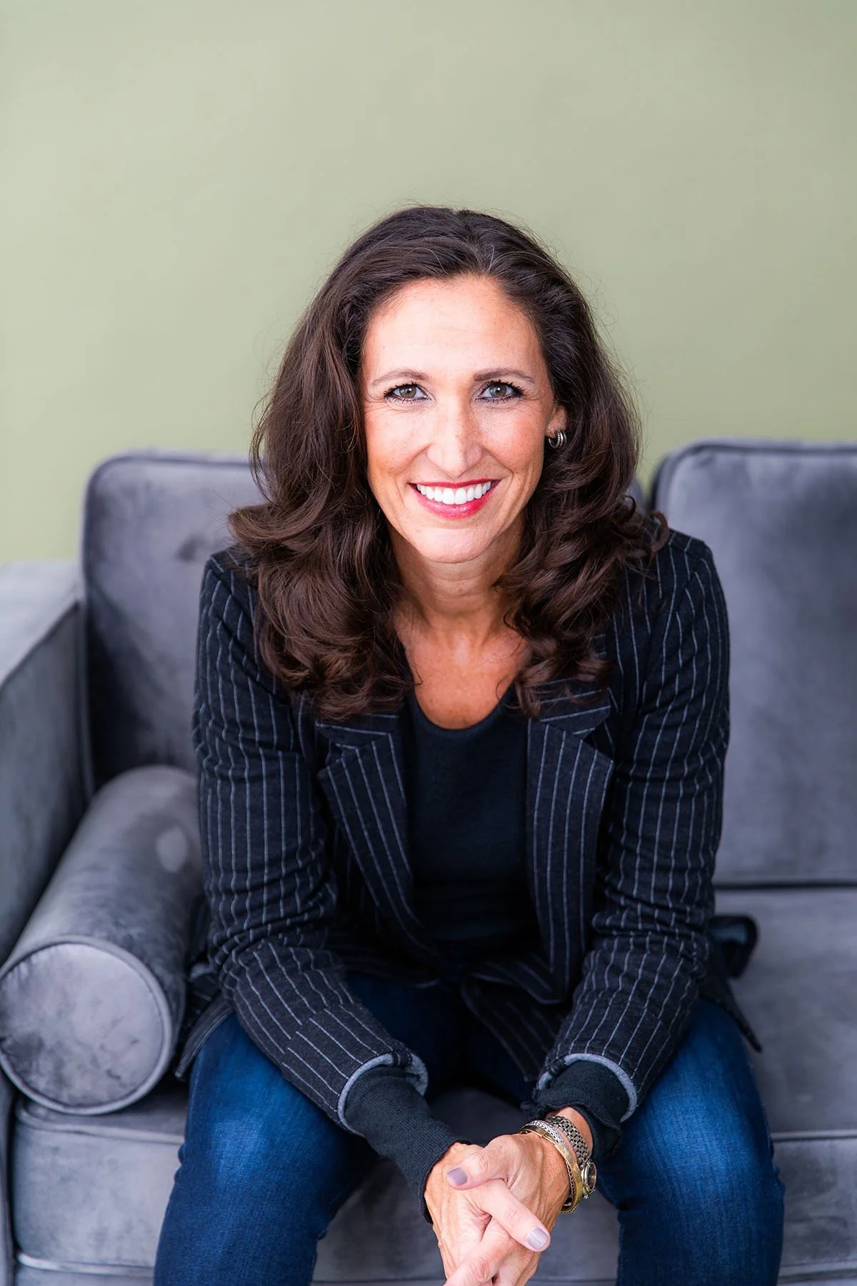 A woman with long, wavy dark hair, wearing a black top and a striped blazer, sitting on a gray couch with her hands clasped. She is smiling and looking directly at the camera.