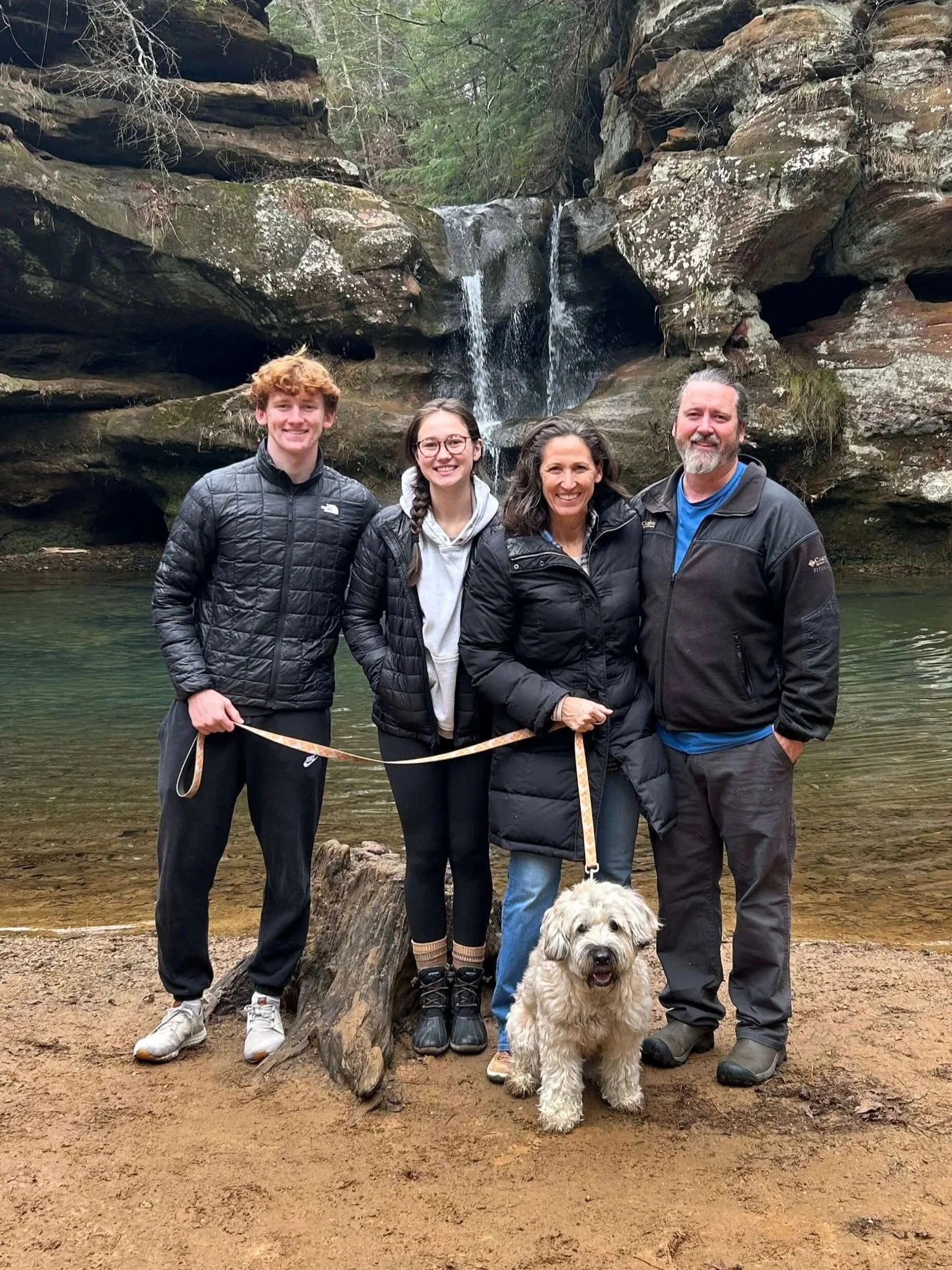 Four people and a dog standing in front of a waterfall in a forest, with rocky cliffs and trees in the background.