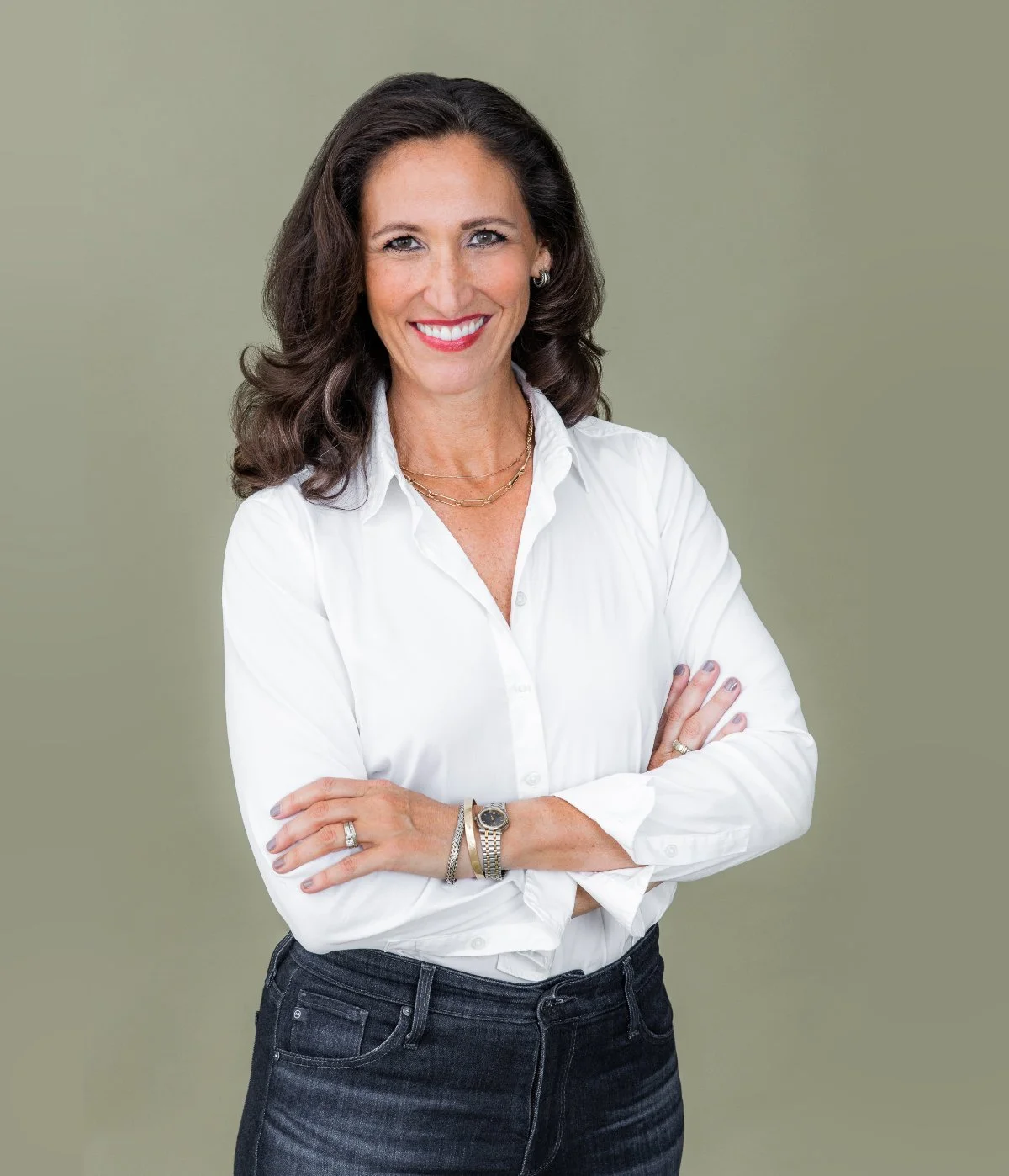 A woman with dark, wavy hair smiling, wearing a white button-up shirt with arms crossed, accessorized with jewelry, standing against a plain, muted green background.