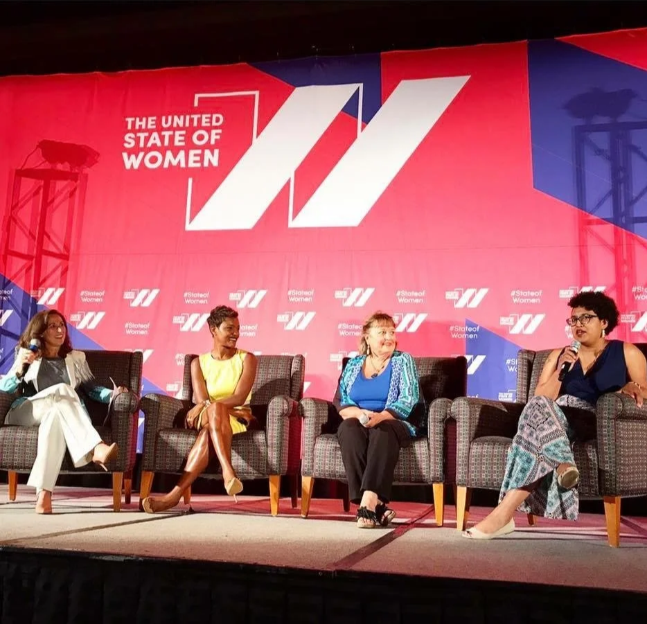 Four women sitting on stage in front of a large banner that reads 'The United State of Women' with the logo and hashtag #StateofWomen. One woman is speaking into a microphone, and the other three women are listening.