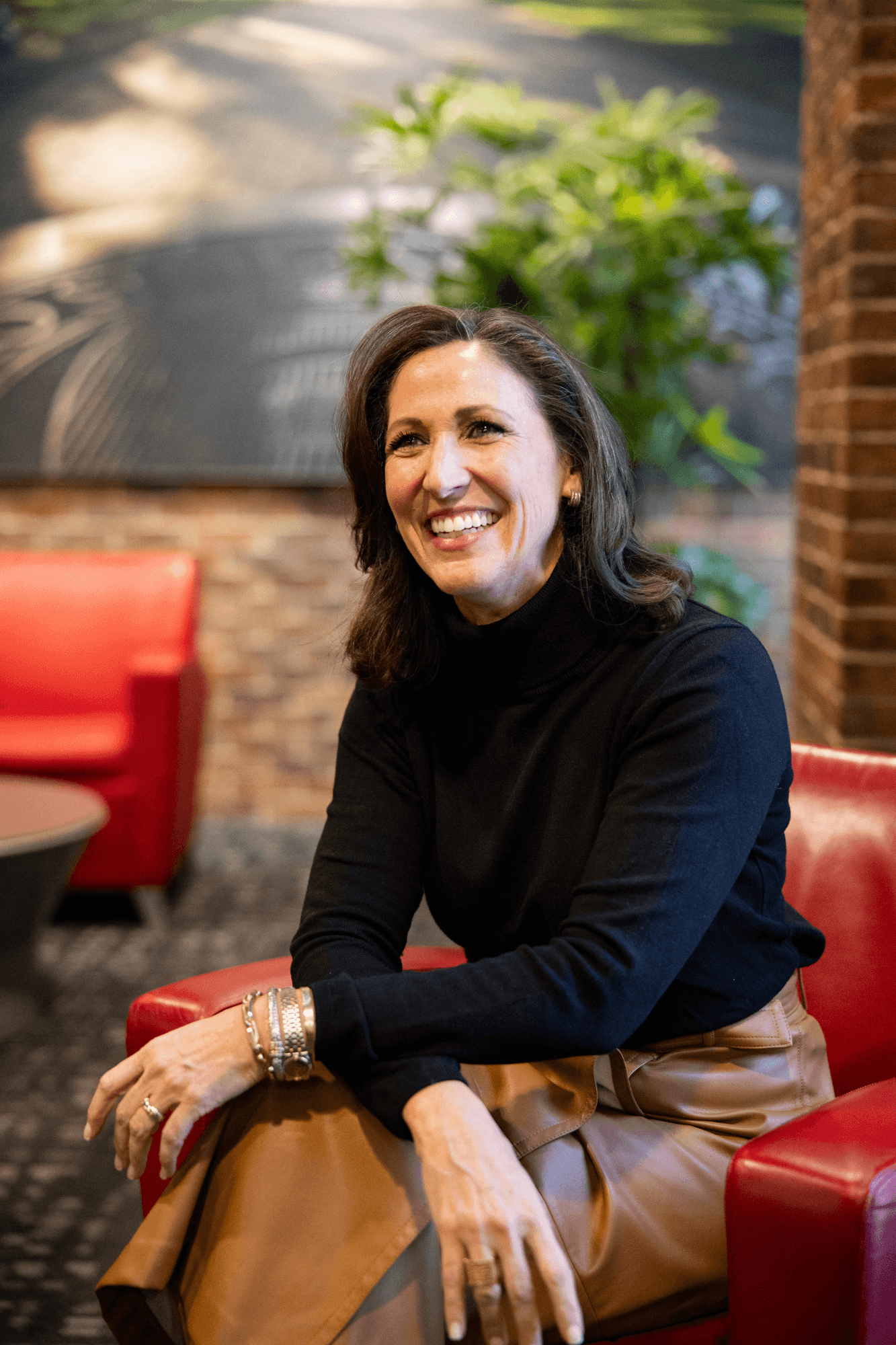 A woman with dark hair smiling, sitting on a red chair in a warmly lit indoor space with brick walls and a plant in the background, wearing a black turtleneck and tan pants.