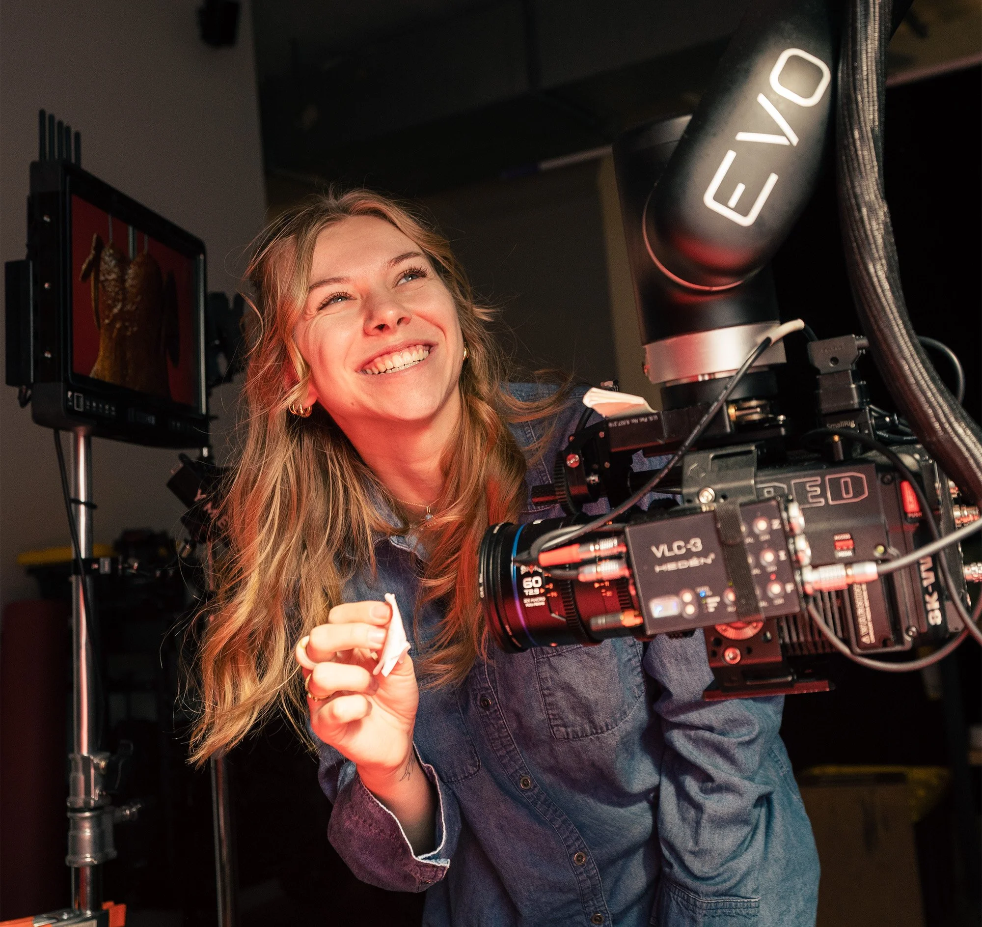 A woman smiling and looking upward while operating a professional film or video camera with a monitor attached. She is in a studio setting.