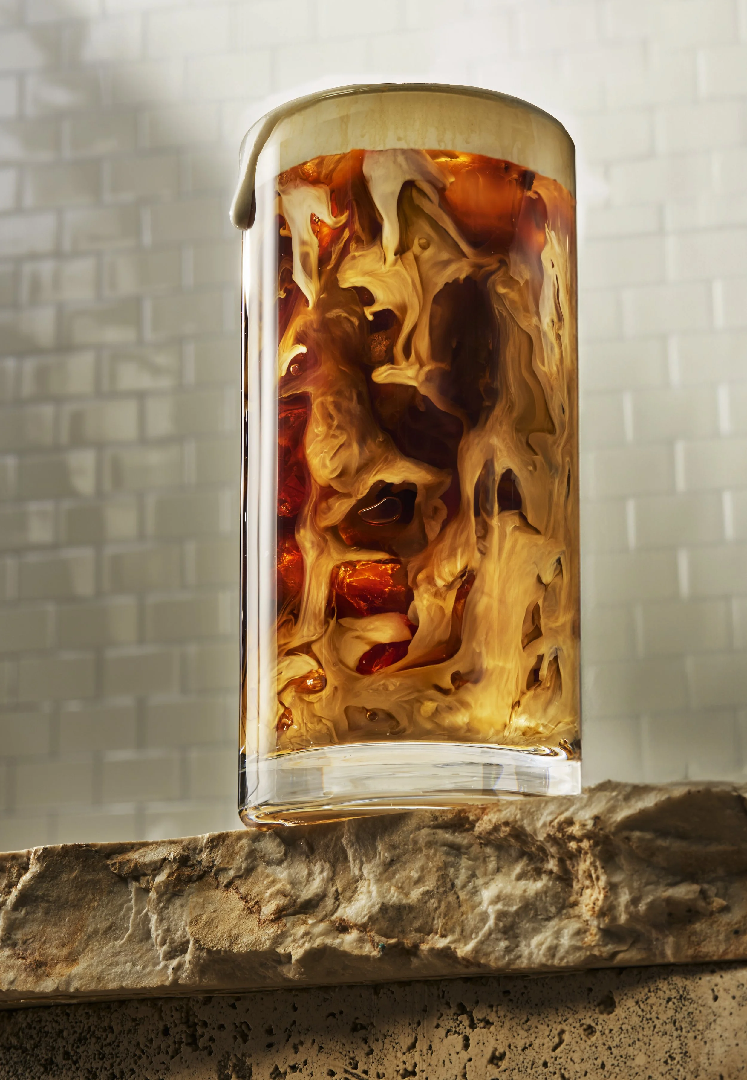 A rectangular glass vase with swirling amber, cream, and brown colors, resembling a marble pattern, set on a textured stone surface against a neutral wall background.