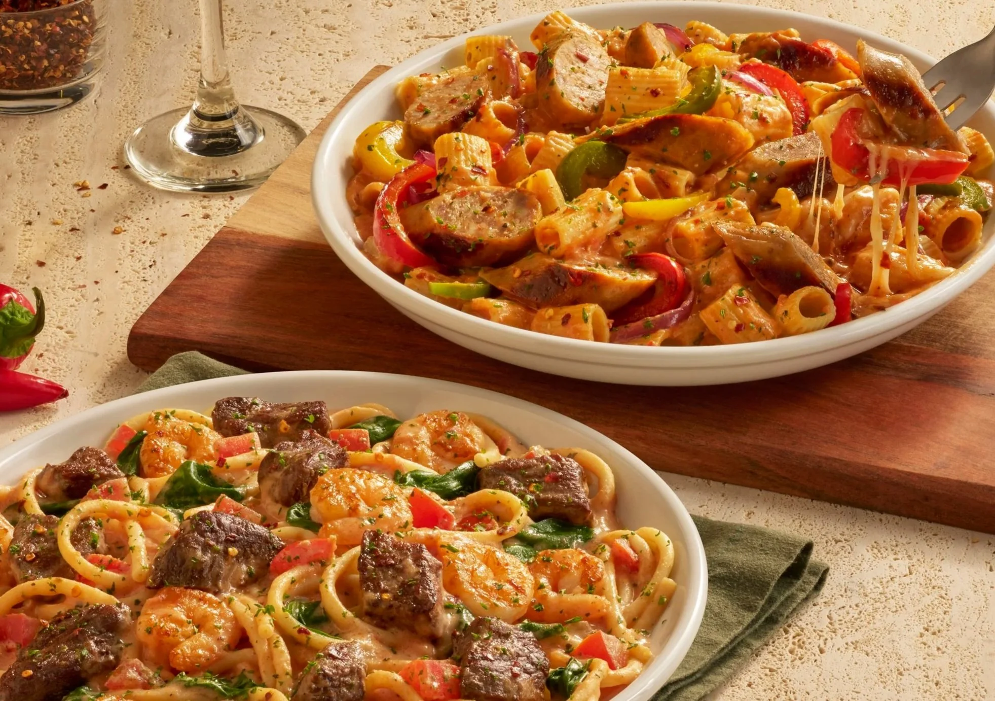 Two white bowls of pasta with sausage, vegetables, and cheese, placed on a dark cloth mat on a textured surface. A red chili pepper and a glass of red pepper flakes are nearby.