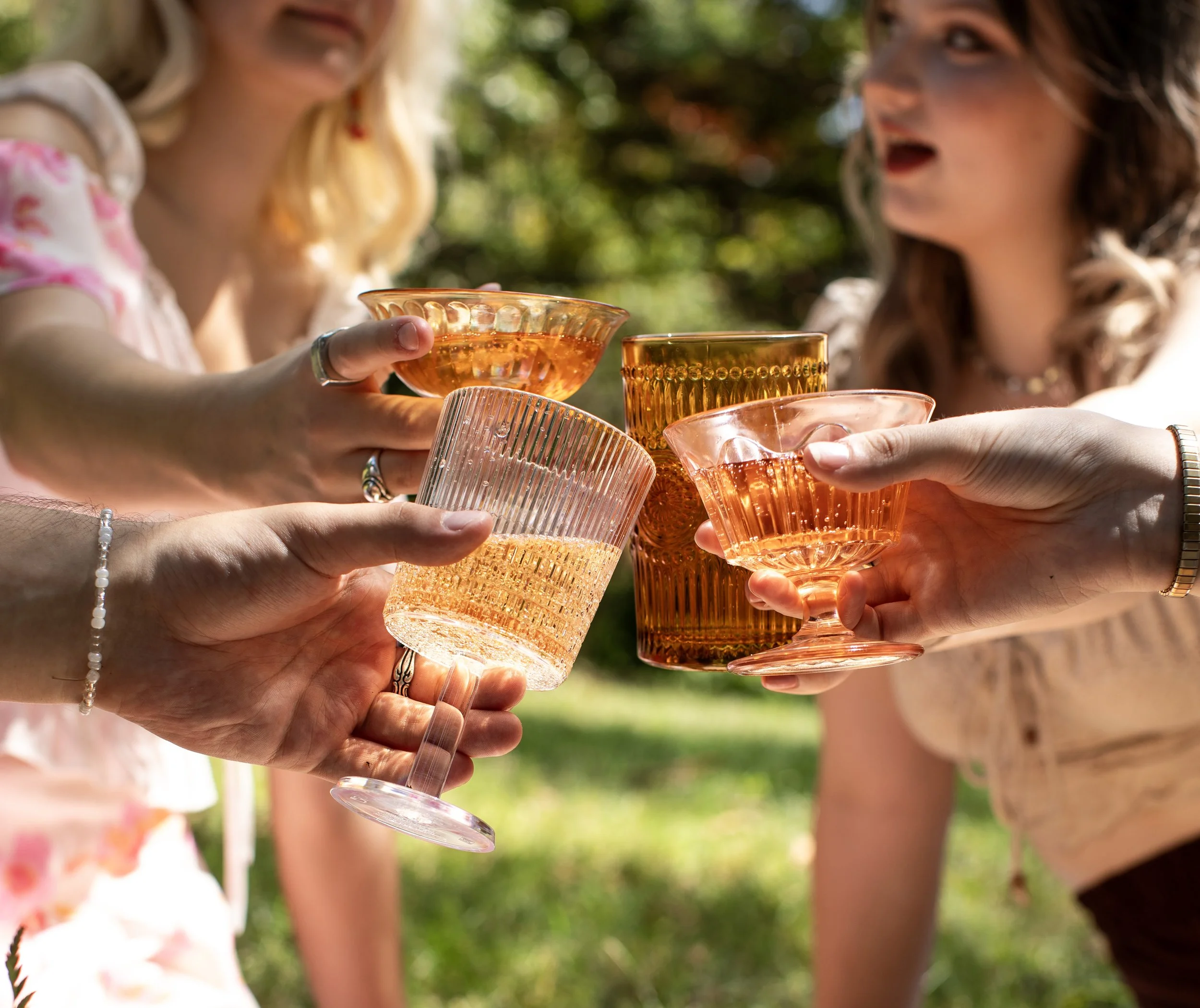 Close-up of multiple hands holding colorful glass cups in an outdoor setting, with blurred green trees in the background.