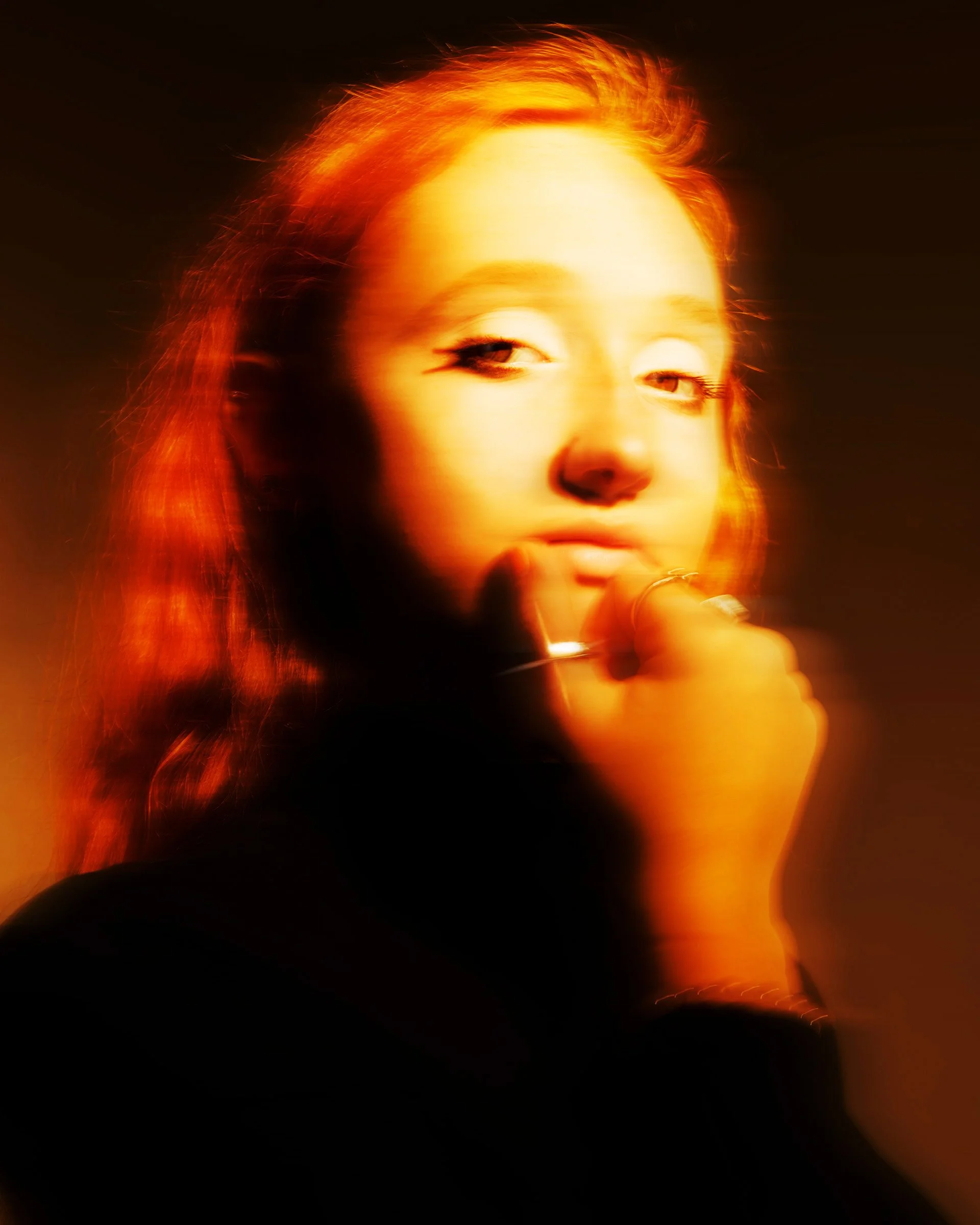 A woman with reddish hair and light skin holding a cigarette close to her lips, illuminated by a warm, orange glow, with a dark background.