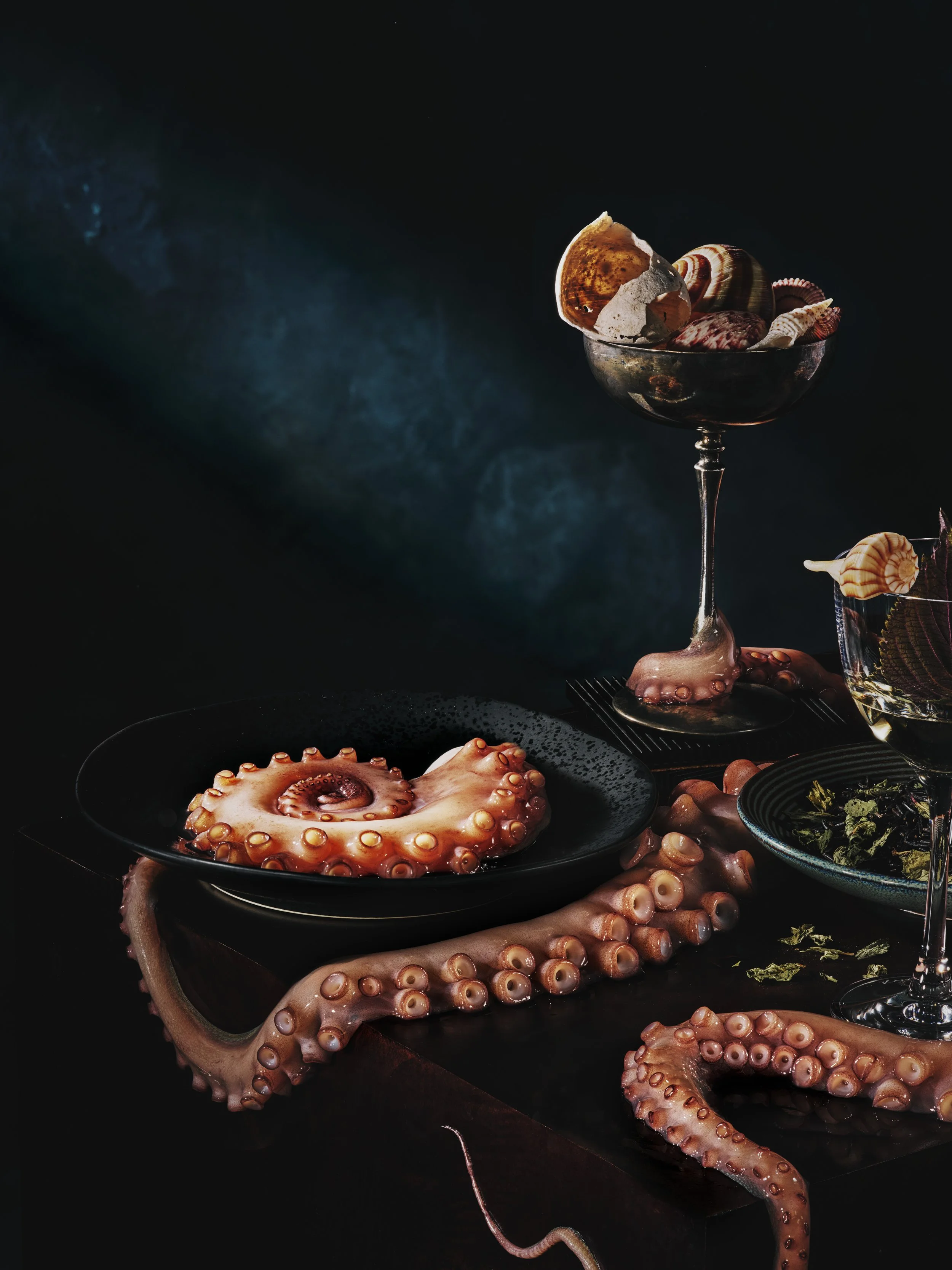 A dark scene with a glass bowl filled with assorted seashells, including a large octopus tentacle with suckers, scattered around the base of the bowl, and additional smaller dishes containing seafood and greens.
