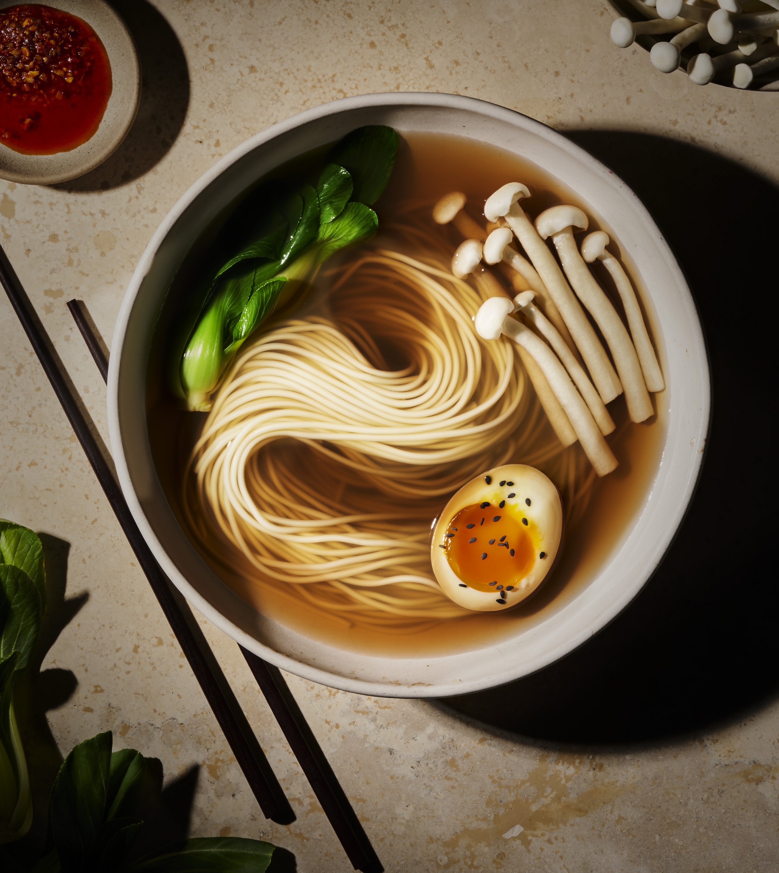 A bowl of ramen noodles with a soft-boiled egg, green leafy vegetables, and enoki mushrooms in a broth. A small bowl of chili flakes or spice is beside the main dish, and a pair of chopsticks rests on the side.