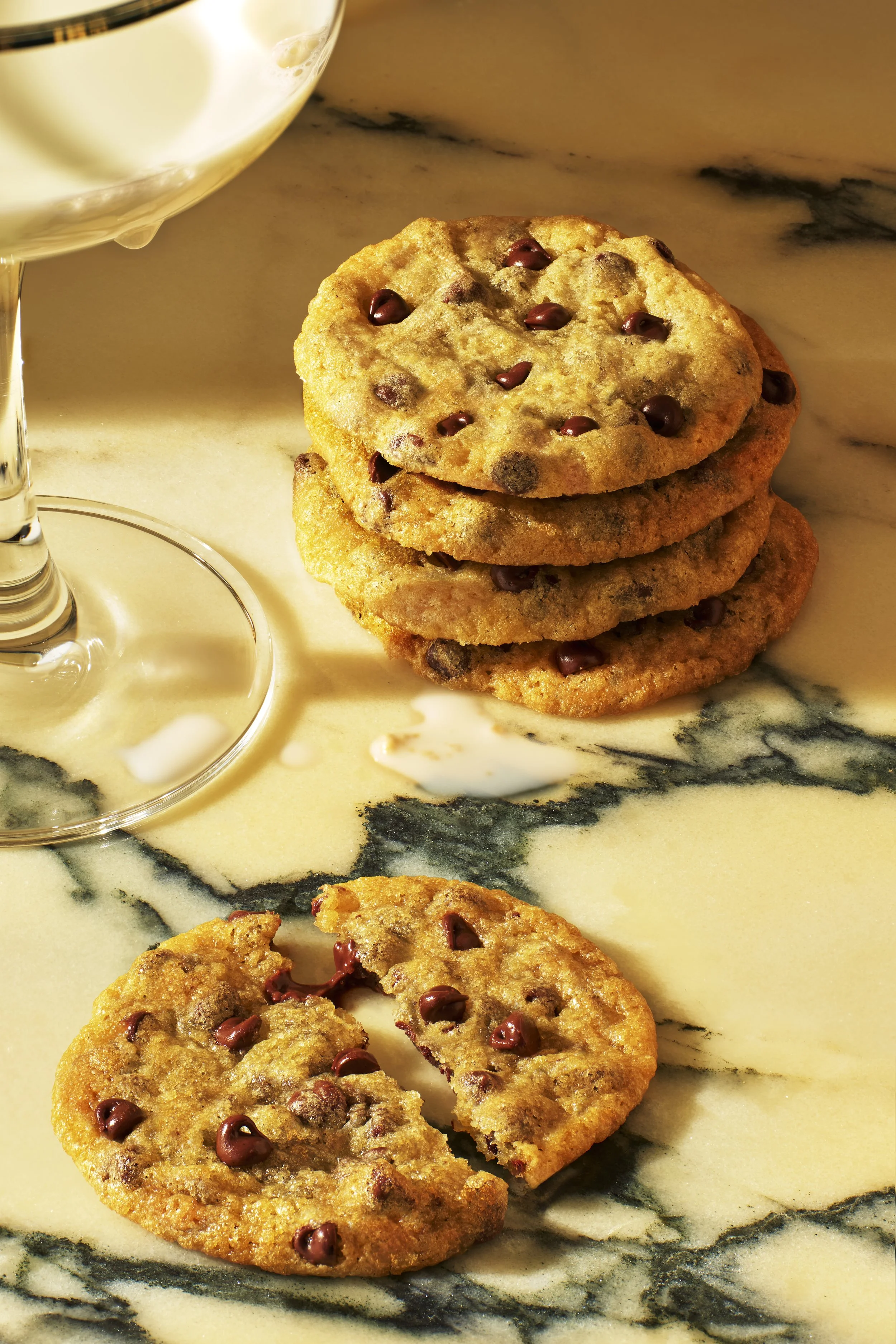 Stack of four chocolate chip cookies and one broken cookie on marble surface with white wine in glass.