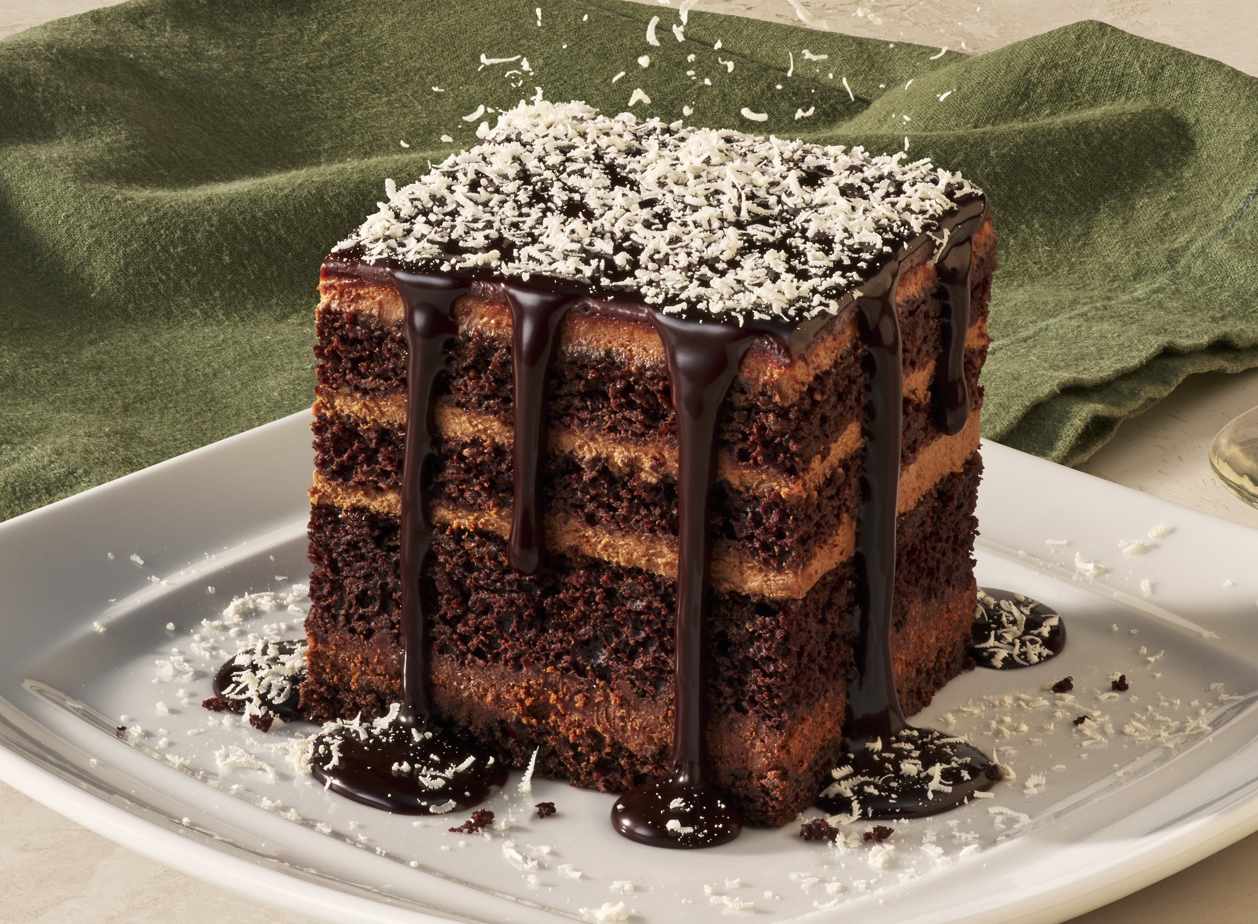 A slice of chocolate cake with chocolate ganache and white shredded topping on a white plate, with dark green cloth in the background.
