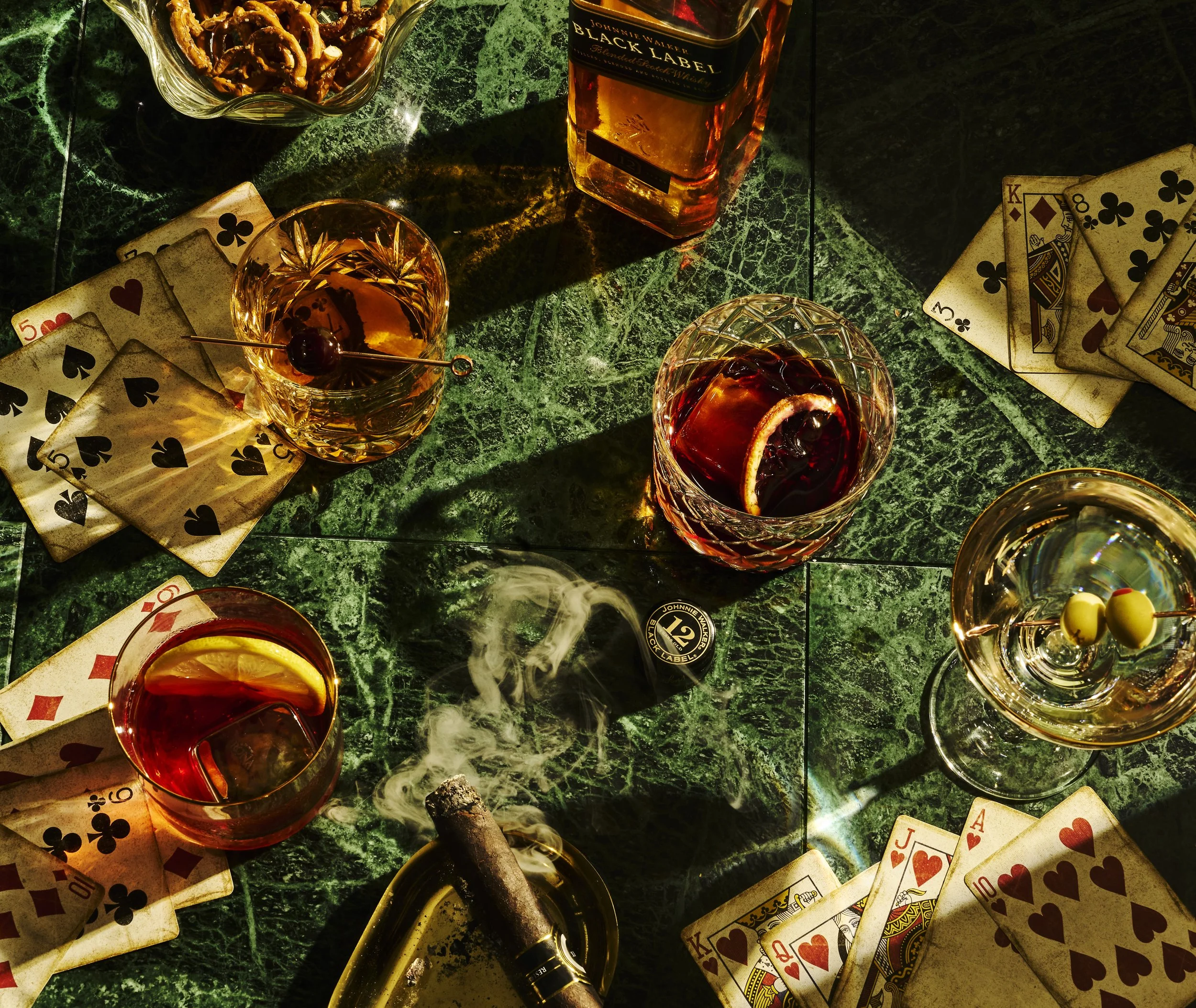 A green marble table with scattered playing cards, glass of whiskey with a cherry, bottle of Jack Daniel's whiskey, ashtray with a cigar, and a glass of whiskey with a cherry and lemon slice.