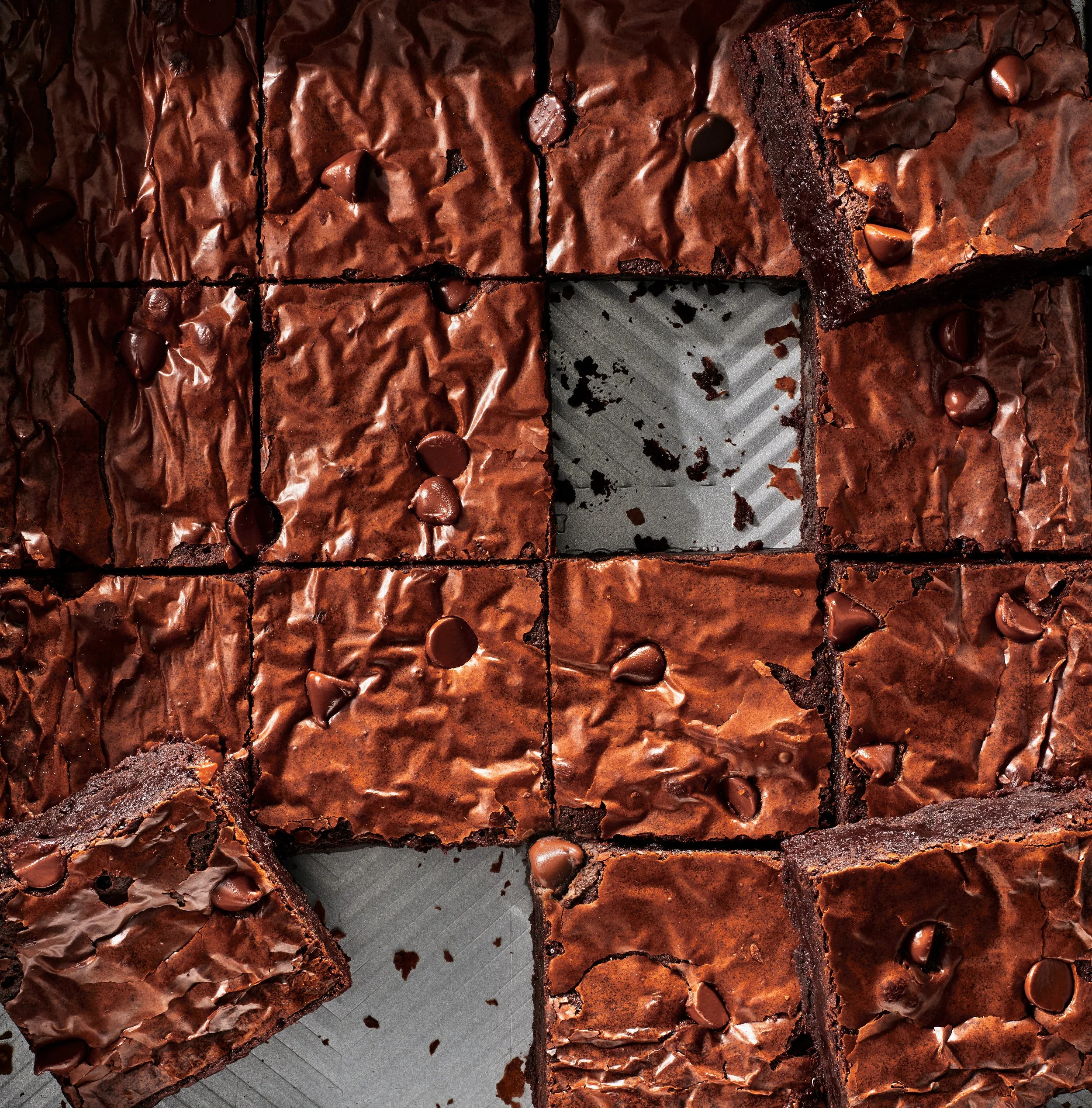 Several pieces of chocolate brownie dessert topped with caramelized chocolate chips and drizzled with chocolate, arranged on a gray surface with one piece cut out.