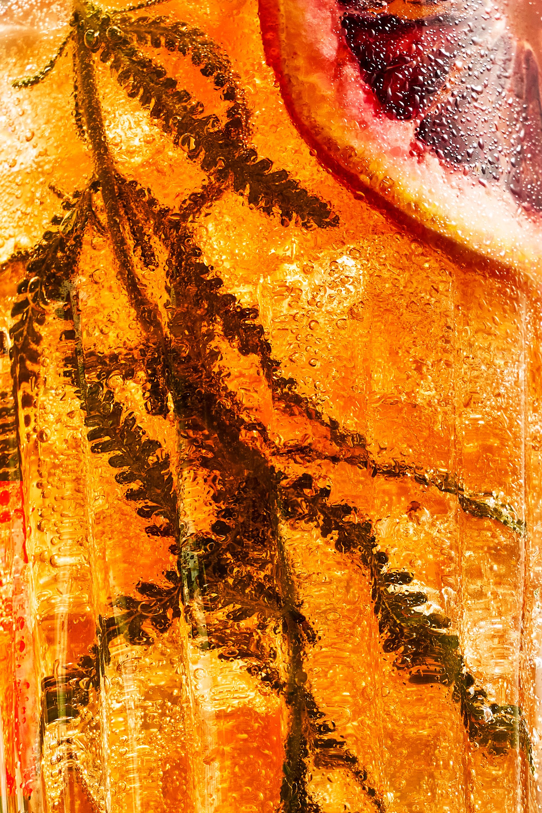 Close-up of a beverage with ice, lemon slices, rosemary, and cherry slices inside, covered in condensation.