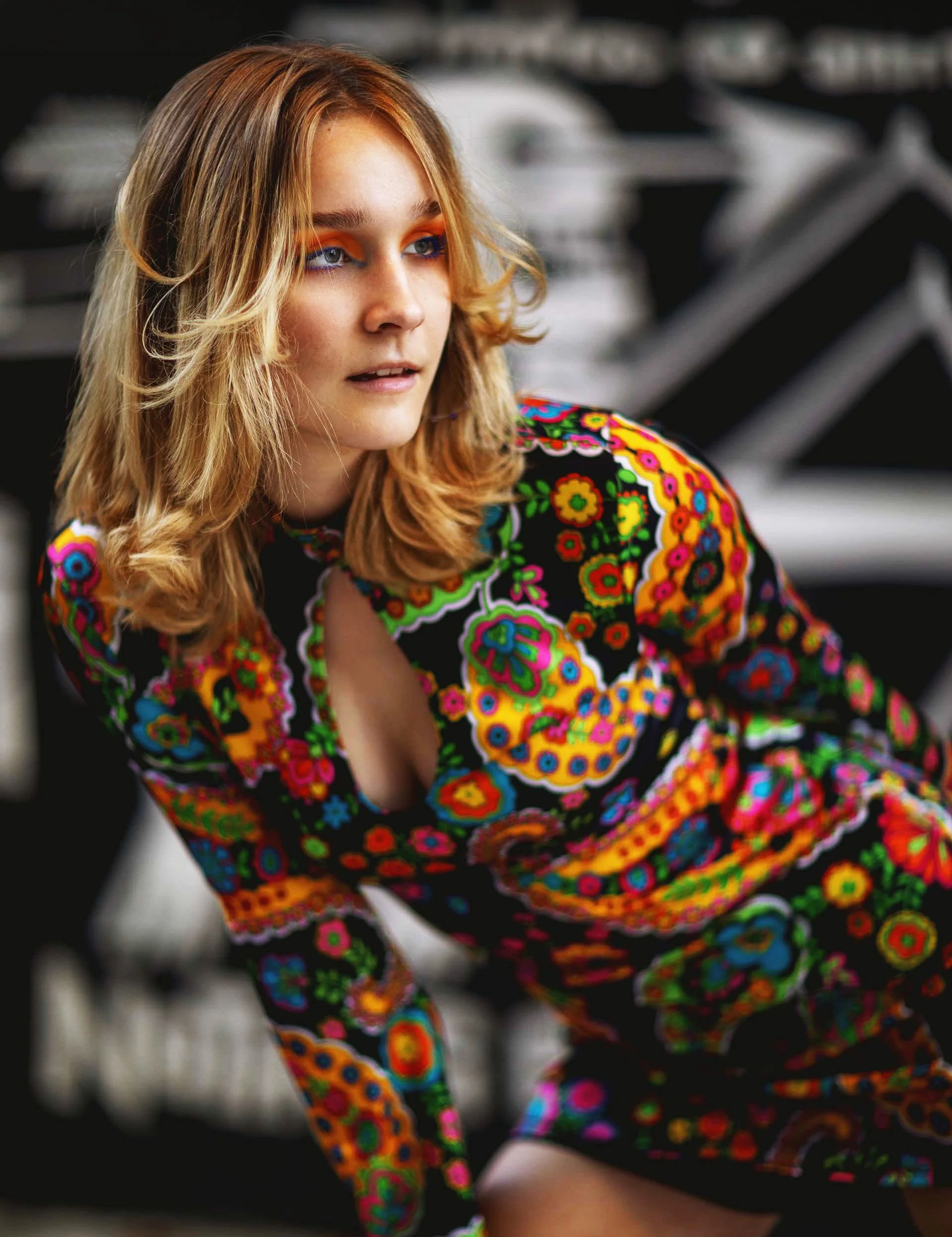 A young woman with wavy red hair and blue eyes looking to the side, wearing vibrant black dress with colorful embroidered floral patterns, in a blurred indoor setting.