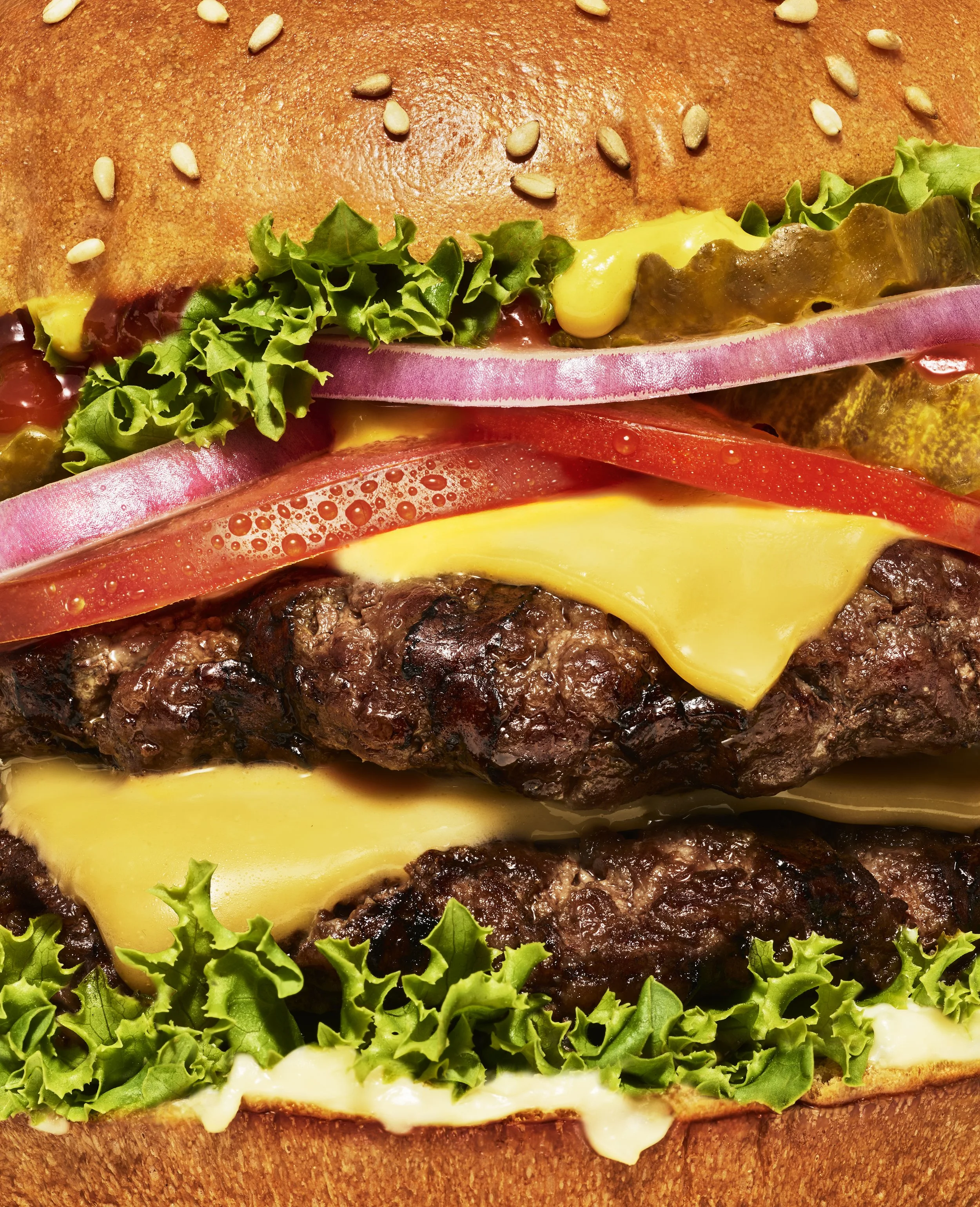 Close-up of a cheeseburger with lettuce, tomato, onion, pickles, melted cheese, and a sesame seed bun.