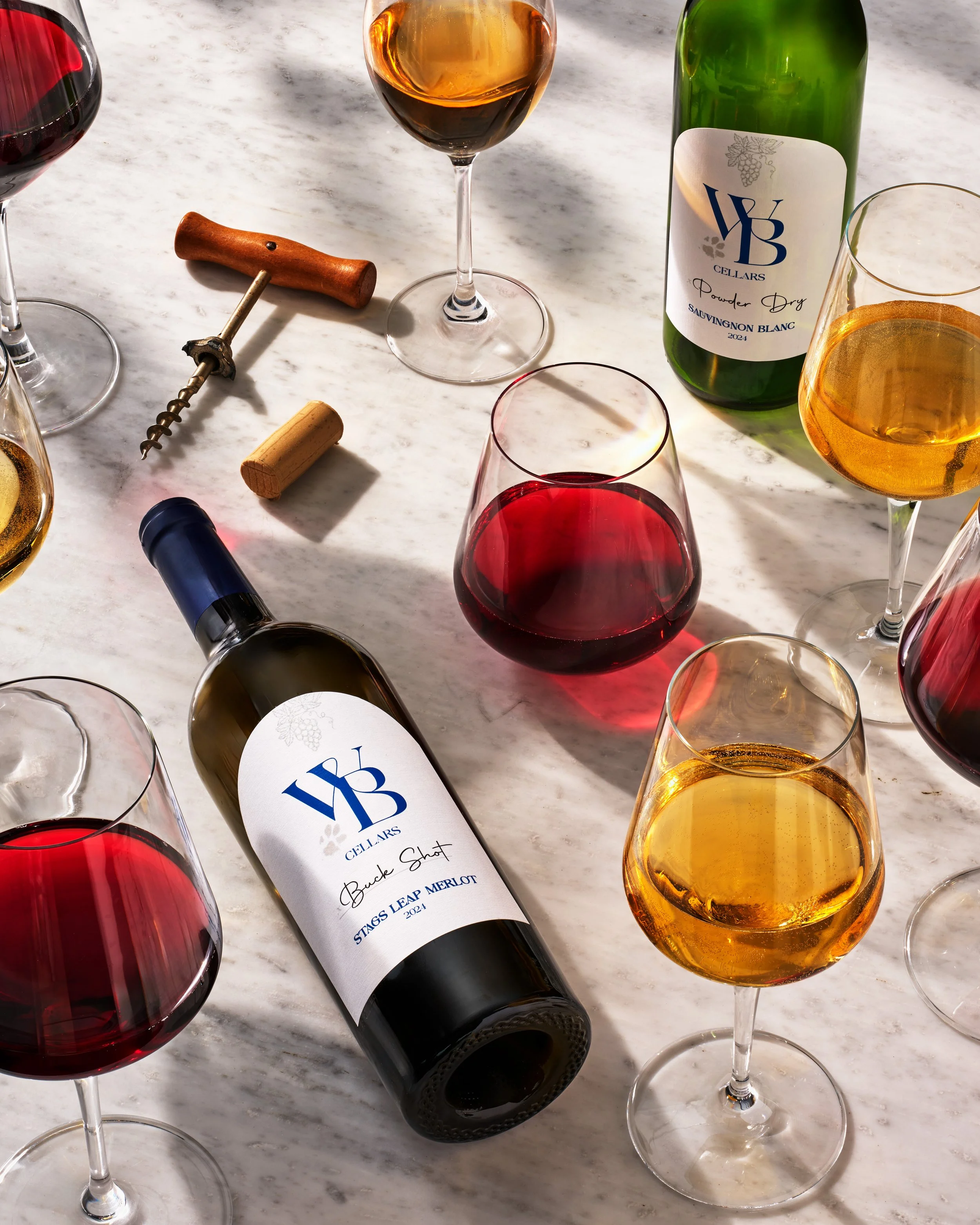 Multiple glasses of red, white, and rosé wine on a marble surface, with a bottle of white wine labeled 'YB Cellars' and a wine cork and corkscrew nearby.