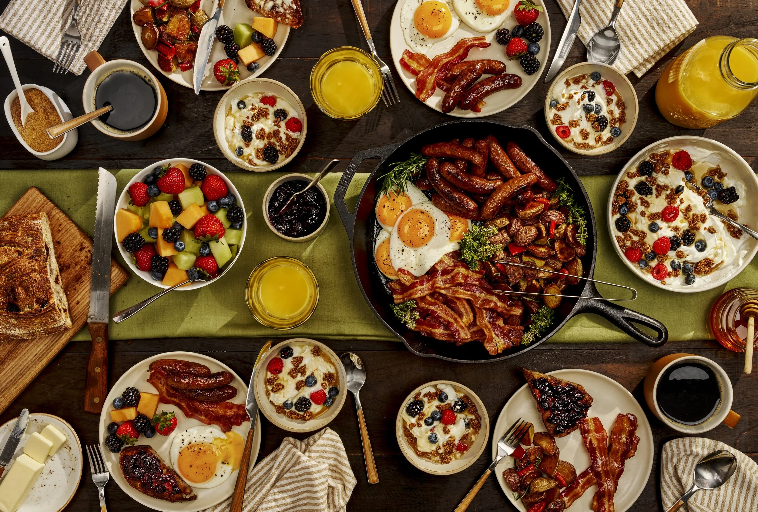 A breakfast spread on a dark wooden table featuring fried eggs, bacon, sausages, fresh fruit, yogurt with berries and granola, coffee, orange juice, and toast.