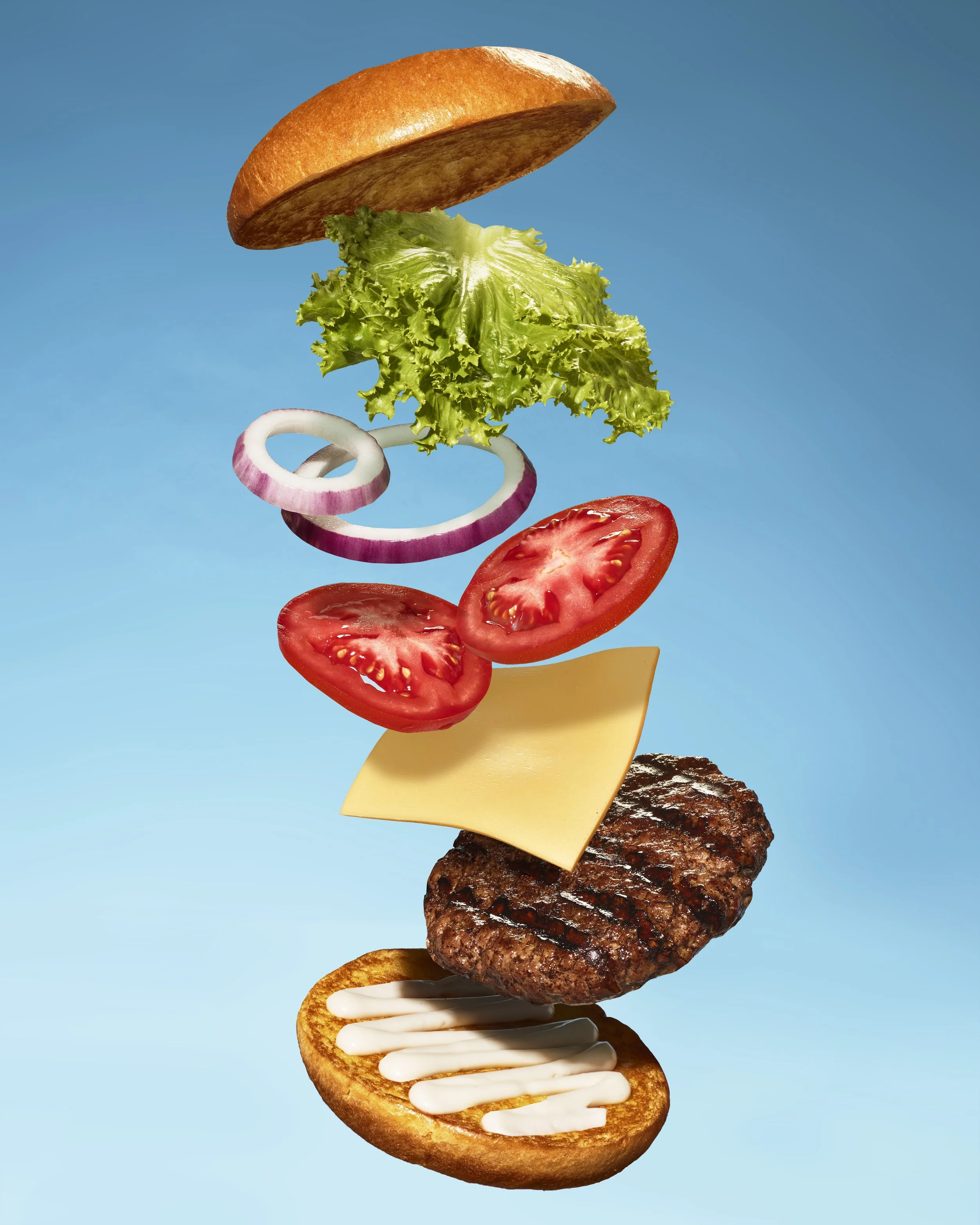 Floating layers of a burger with bun, lettuce, tomato, onion, cheese, hamburger patty, and condiments set against a blue background.