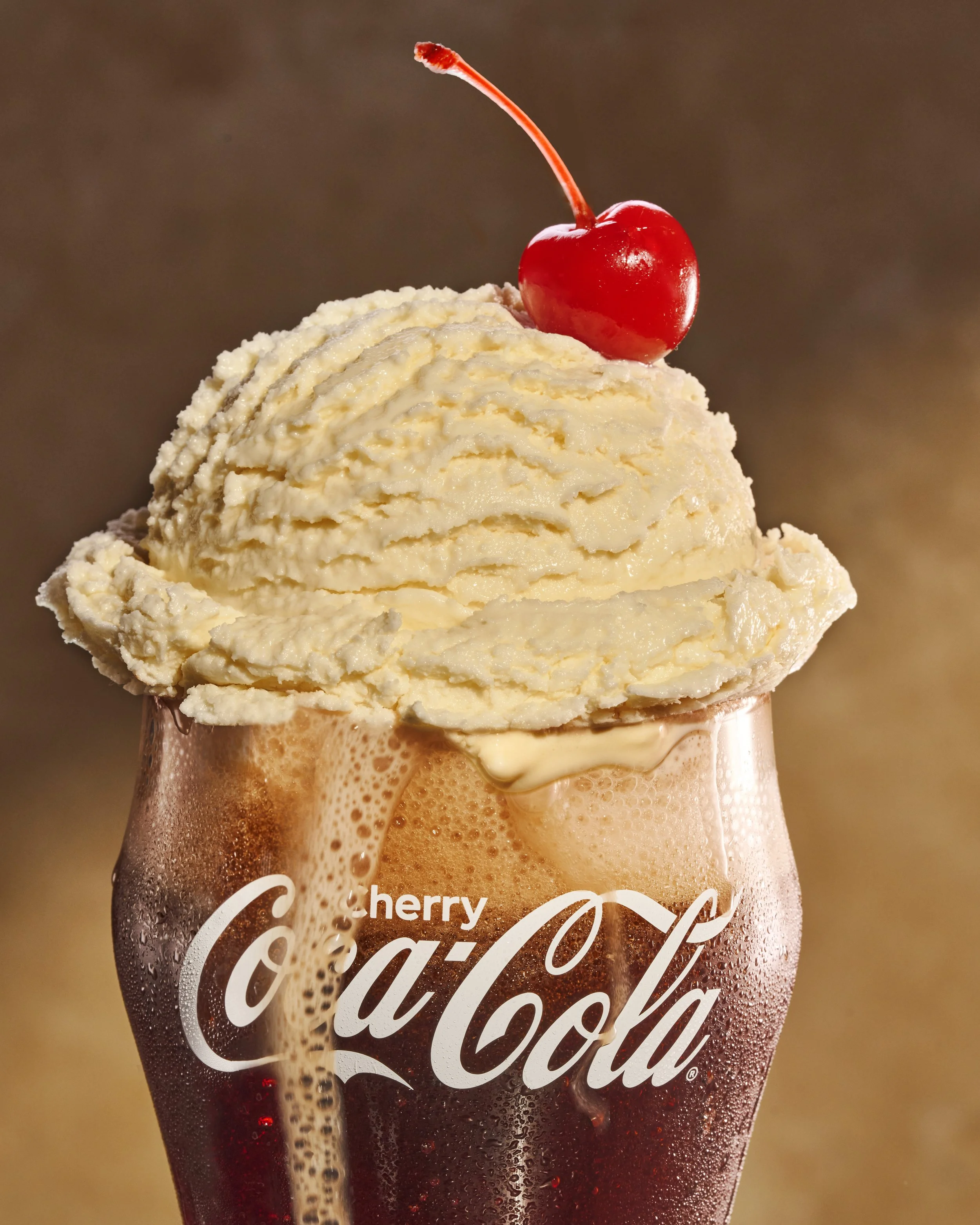 Close-up of a glass of Coca-Cola with ice and a cherry on top, condensation on the glass, and a Coca-Cola logo on the glass.