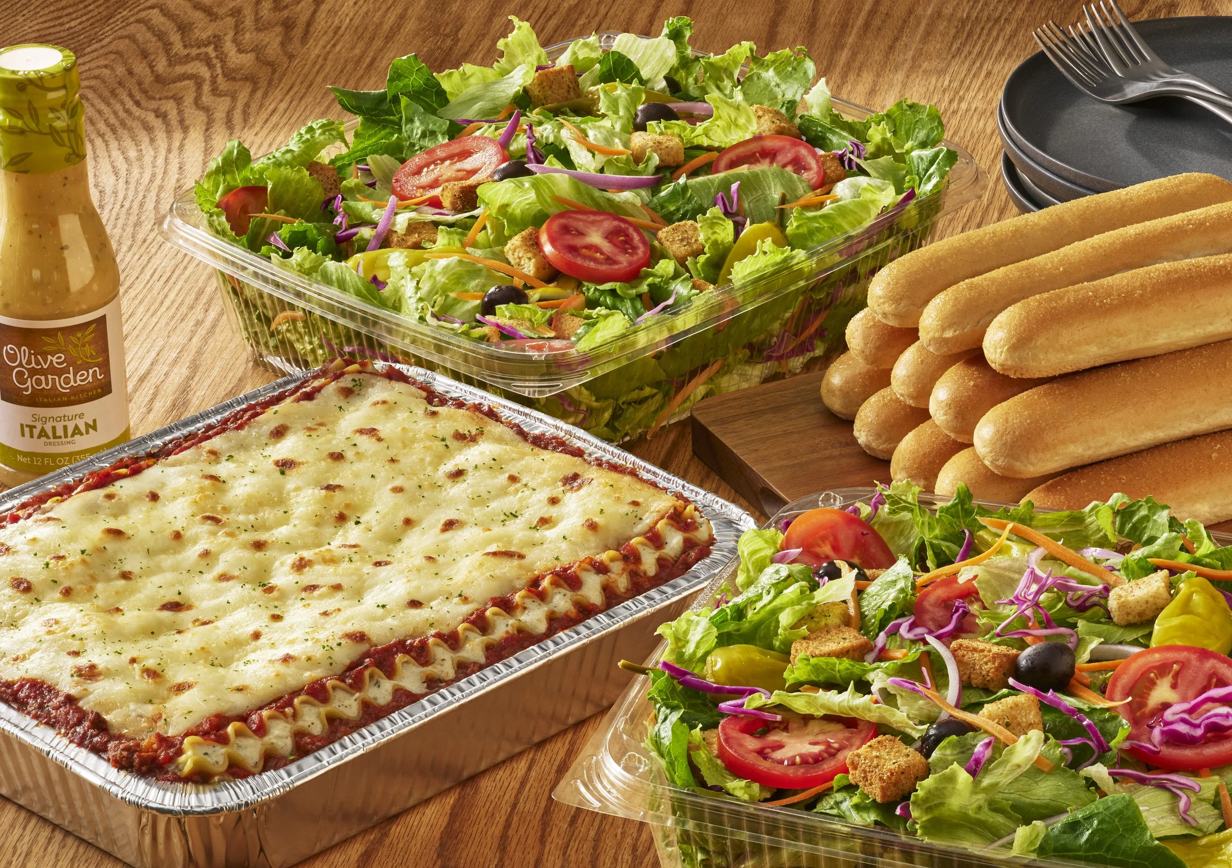 A spread of food on a wooden table, including a large salad with fresh vegetables, a tray of baked cheesy pasta, a bottle of Italian dressing, and a stack of breadsticks.