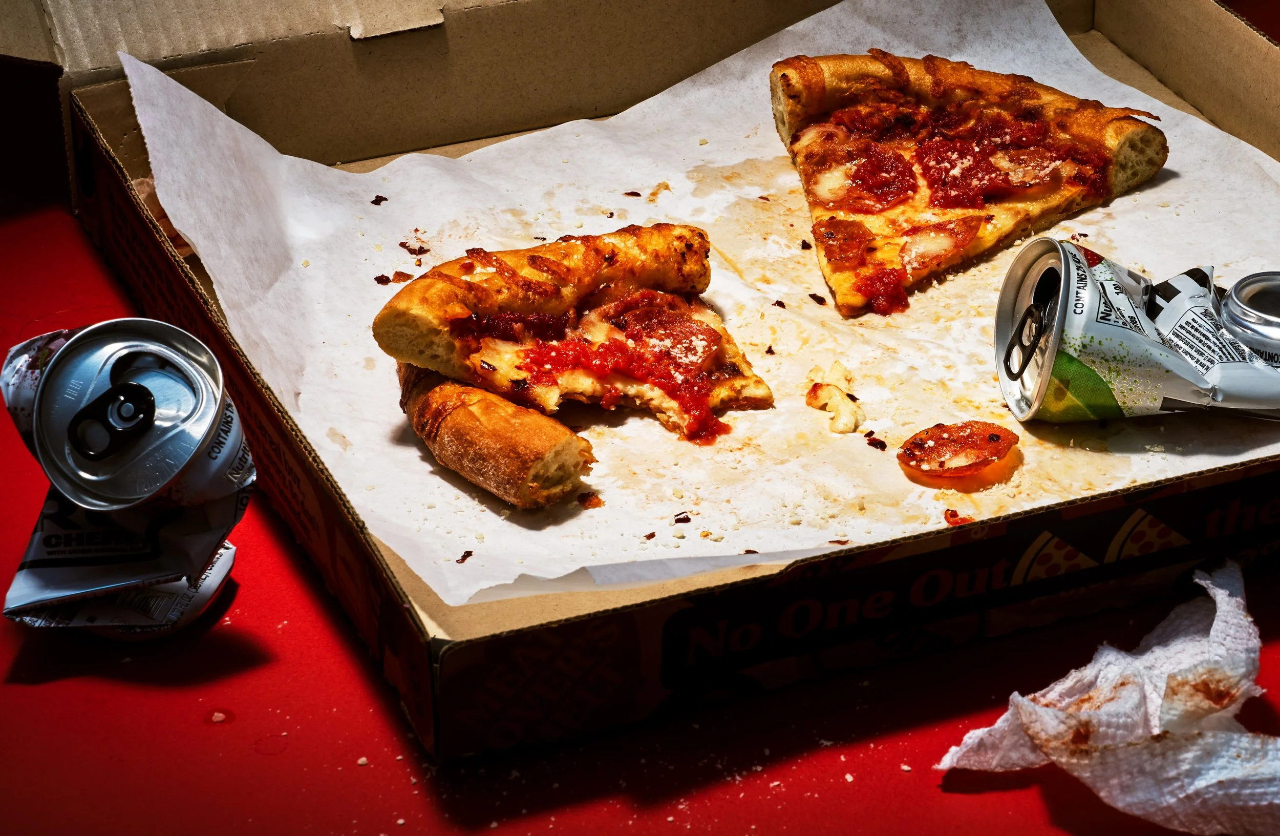 A partially eaten pizza in a box with two empty soda cans on a red surface.