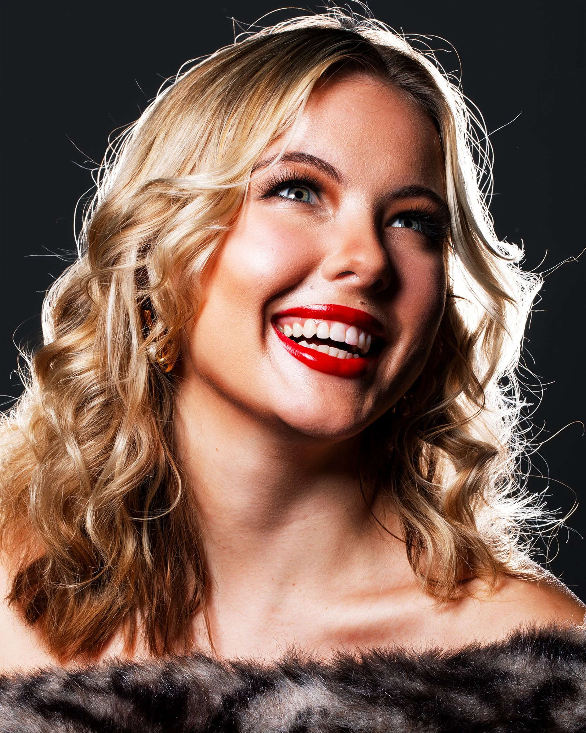 Close-up of a smiling young woman with blonde curly hair, bright red lipstick, and radiant skin, wearing a fuzzy black and gray garment against a dark background.