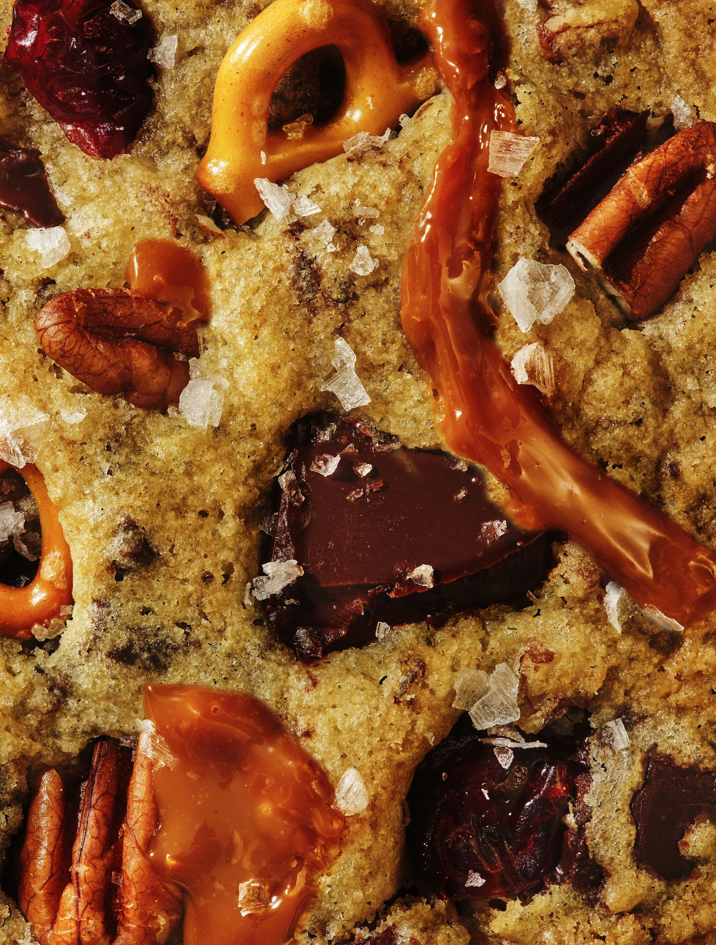 Close-up of a dessert featuring soft, moist cake with caramel or butterscotch sauce, pecan halves, dried cranberries, a dollop of bright orange jam or fruit filling, and coarse sea salt sprinkled on top.
